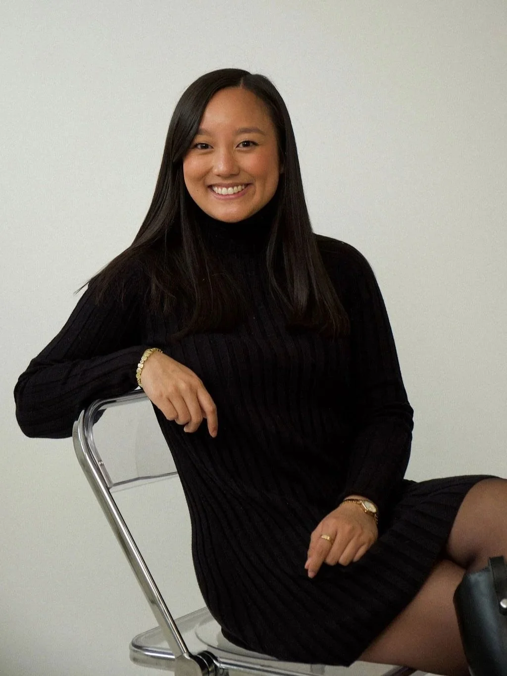 A woman with long black hair wearing a black ribbed dress sitting on a metal chair with a plain white wall background, smiling at the camera.