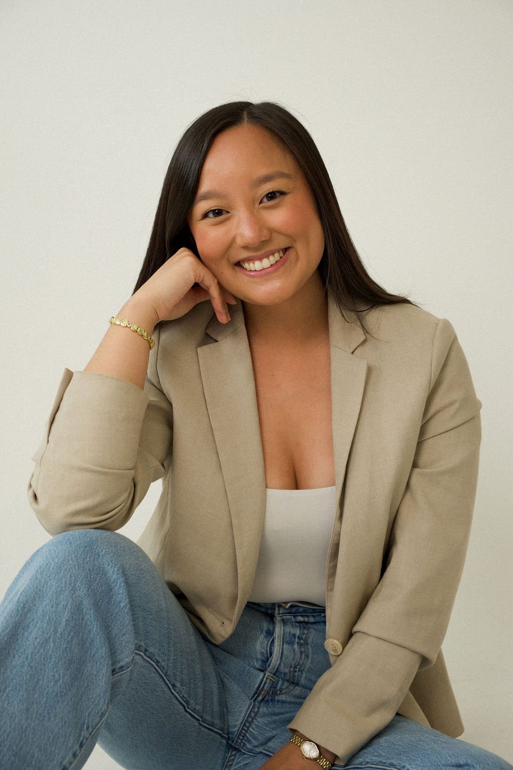 A woman with long dark hair smiling and resting her head on her hand, sitting against a plain white wall, wearing a beige blazer, white top, blue jeans, a gold bracelet, and a watch.