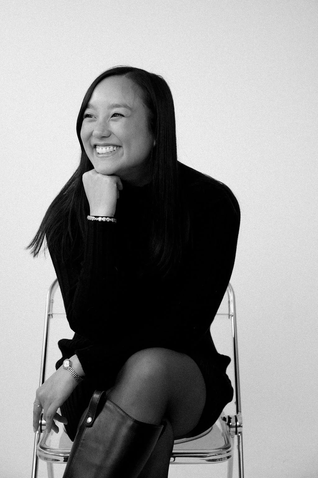 A black-and-white photo of a smiling woman sitting on a chair with her chin resting on her hand, wearing a black long-sleeve top, a bracelet, a watch, and tall boots.