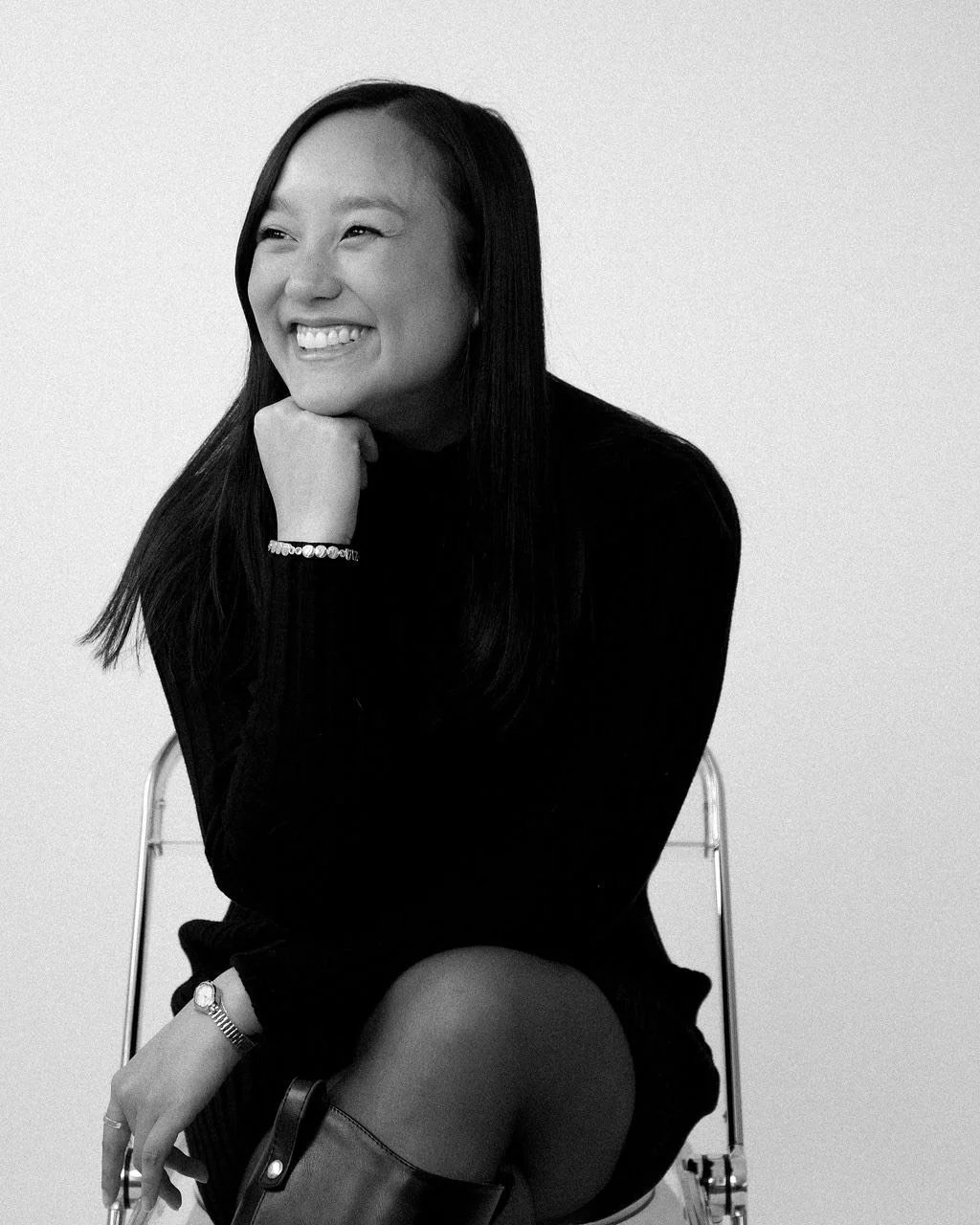 Black and white photo of a woman sitting on a chair, smiling, with her chin resting on her hand, wearing a long-sleeved black top, a bracelet, a watch, and knee-high boots.