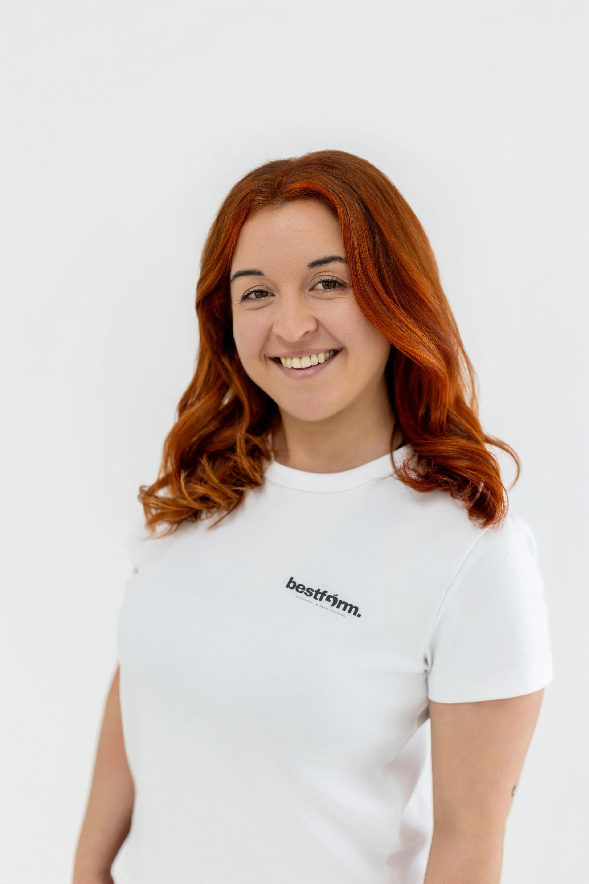 Porträt einer lächelnden Frau mit roten, gewellten Haaren, die ein weißes T-Shirt trägt mit dem Text »bestform.« auf der Brust, vor hellem Hintergrund.