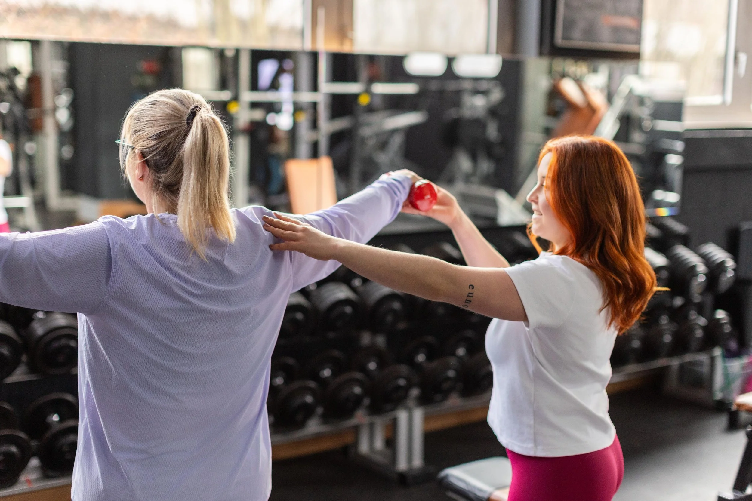 Zwei Frauen trainieren im Fitnessstudio, eine gibt der anderen einen Schubs bei einem Krafttraining, im Hintergrund Hantelbänke.