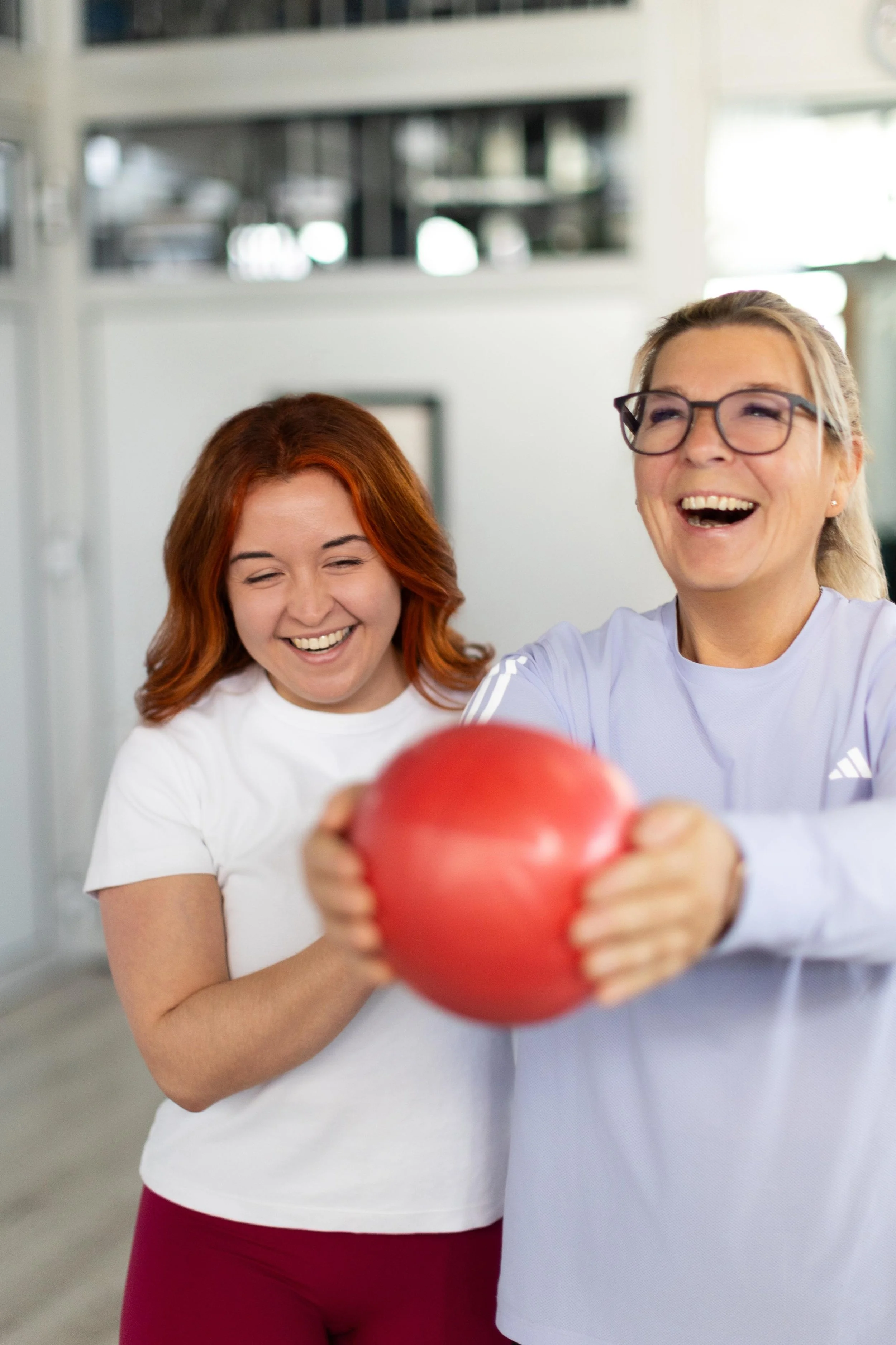 Zwei Frauen, die lachen und einen roten Basketball halten, in einem Raum mit weißen Wänden und Regalen im Hintergrund.