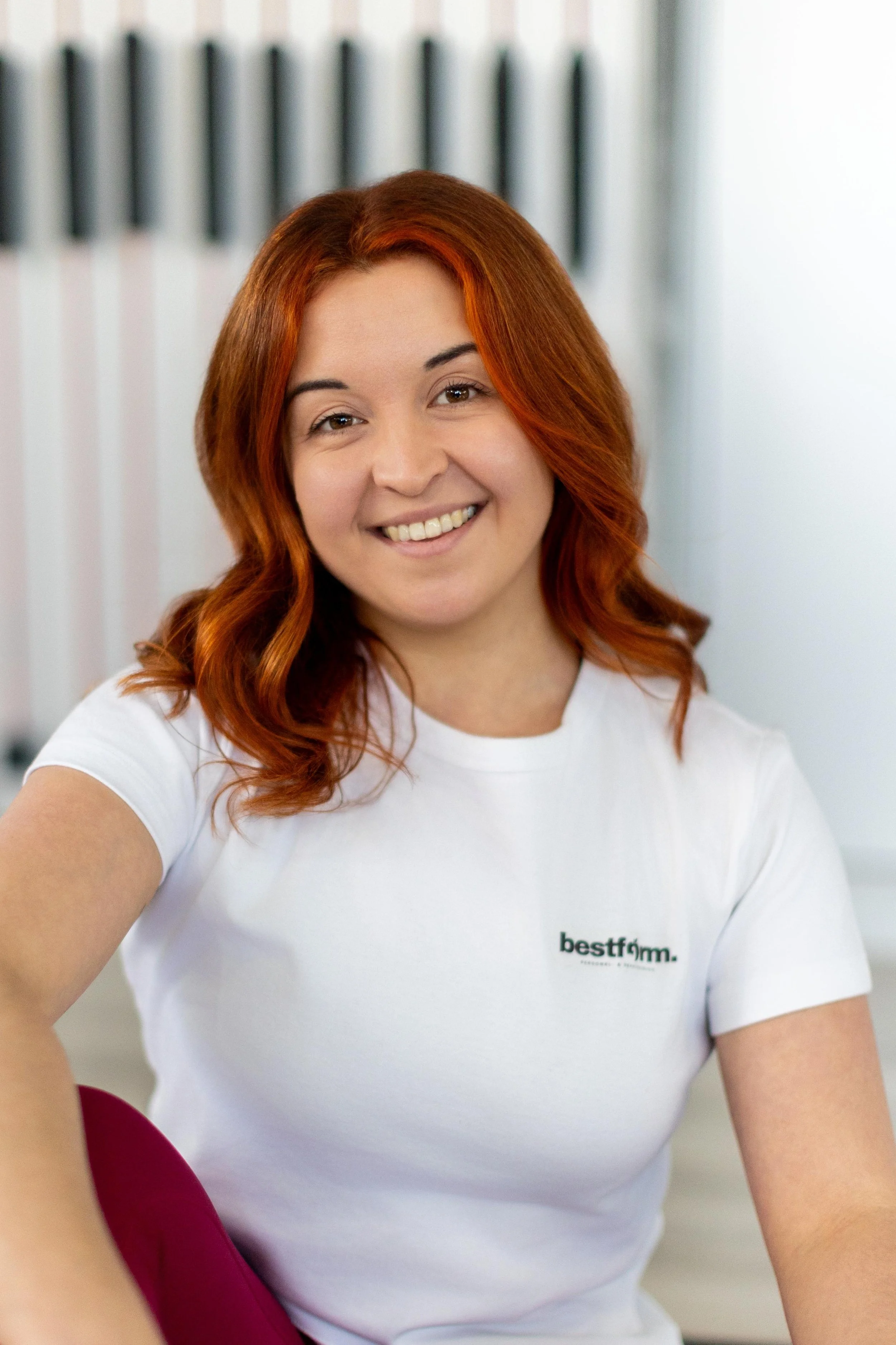 Lächelnde Frau mit roten Haaren, trägt ein weißes T-Shirt mit dem Logo 'bestform', sitzt in einem Raum mit weißen Wänden und einem schwarzen Objekt im Hintergrund.