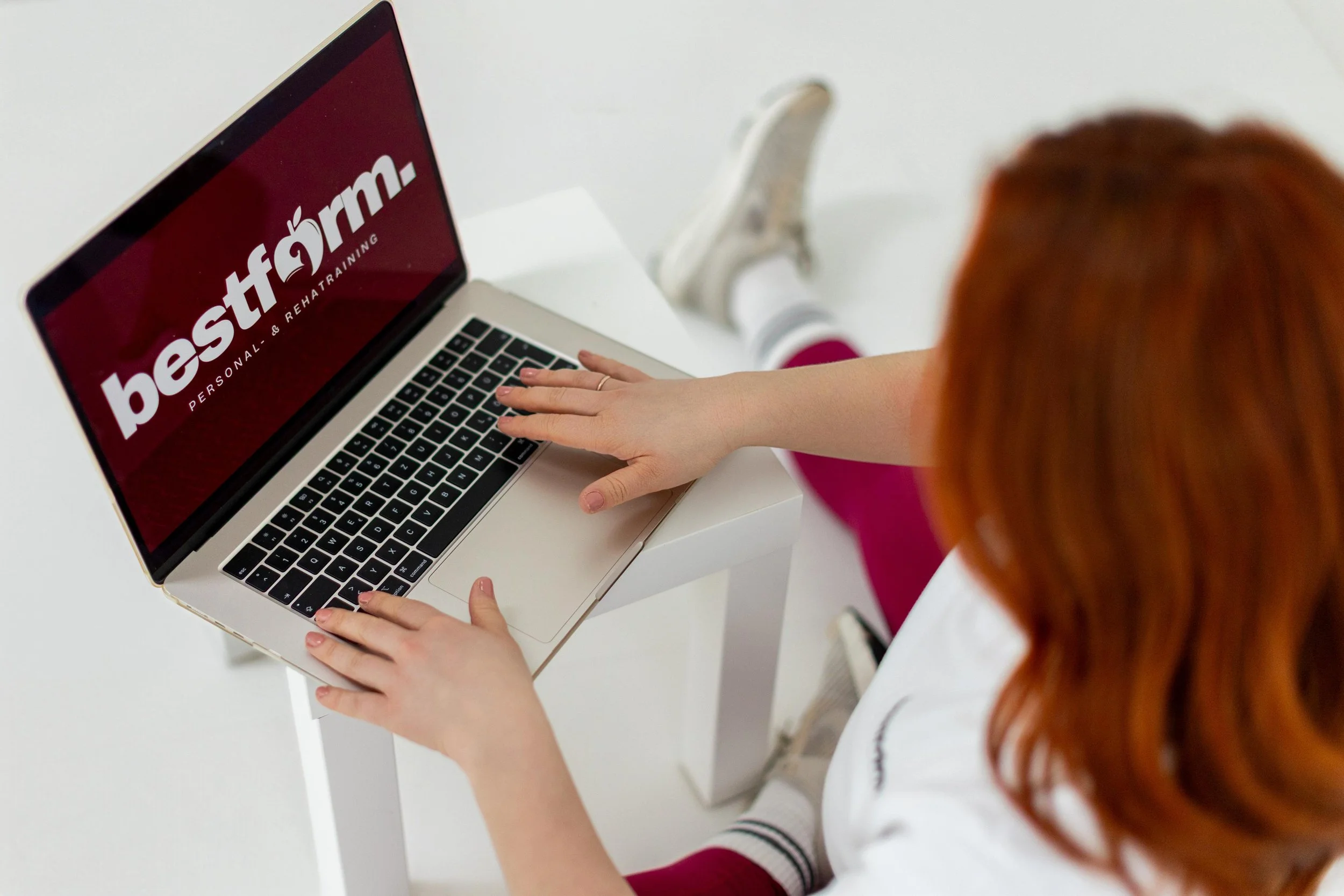 Eine Frau mit roten Haaren nutzt einen Laptop mit einem Bildschirm, auf dem das Logo von 'bestform' mit dem Untertitel 'PERSONAL- & REHA-TRAINING' zu sehen ist, während sie auf einem weißen Tisch sitzt.