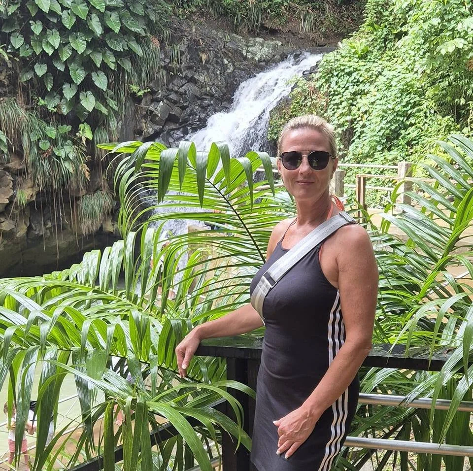 Eine Frau trägt eine schwarze ärmellose Sportkleidung mit weißen Streifen, trägt Sonnenbrille und eine kleine schwarze Tasche, steht vor einem Wasserfall inmitten von grünen Pflanzen im Freien.