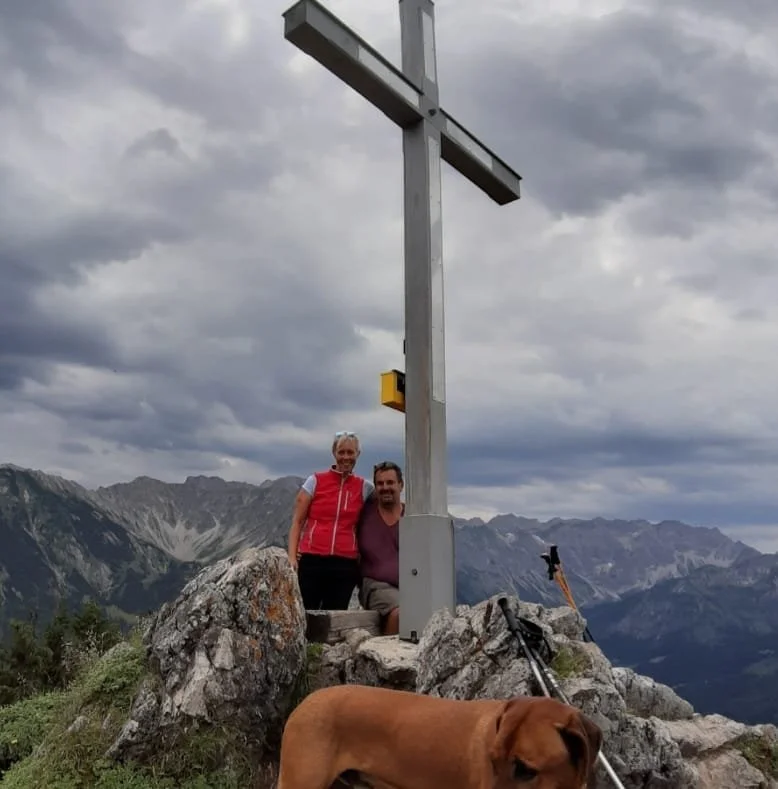 Zwei wandernde Personen vor einem Gipfelkreuz auf einem Berg mit Bergpanorama im Hintergrund, ein Hund liegt im Vordergrund, dunkle Wolken am Himmel.