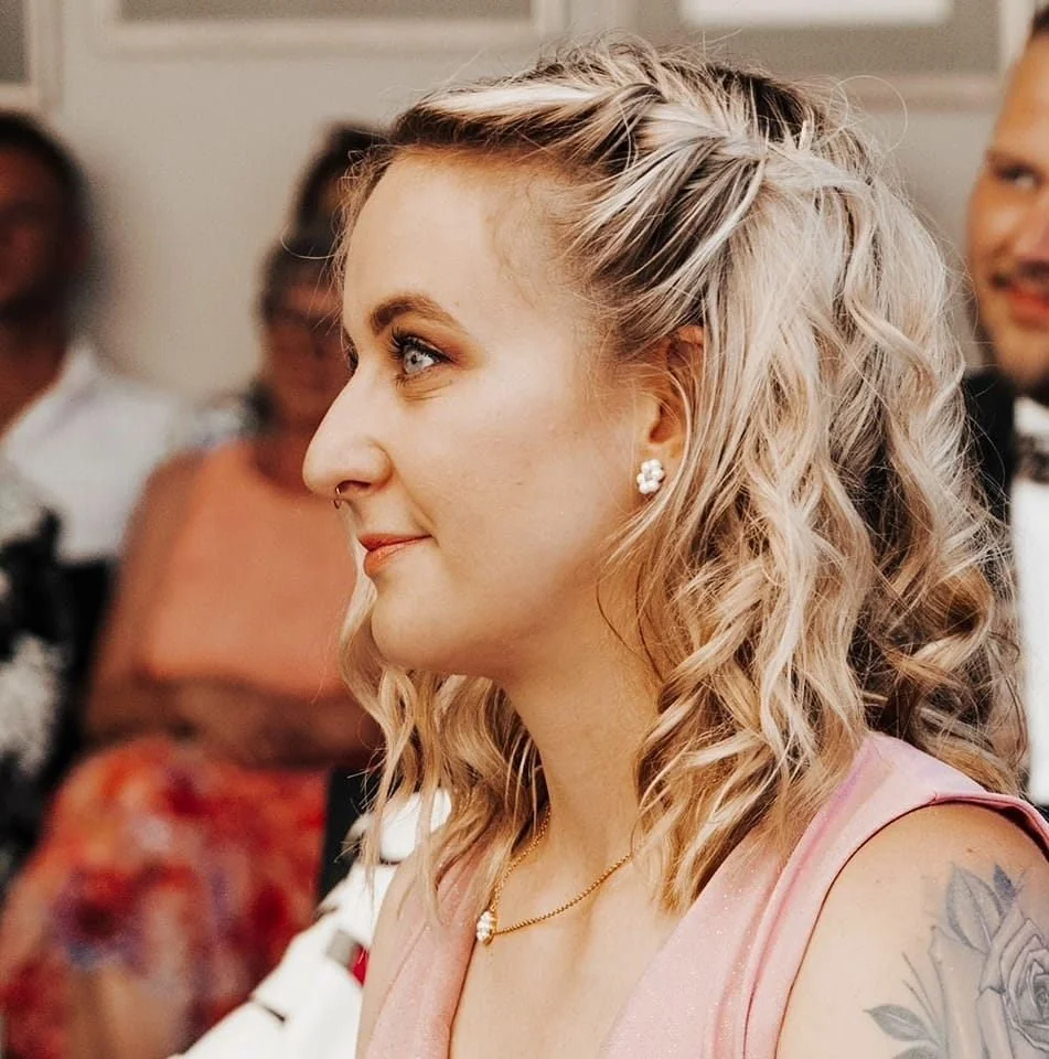 Junge Frau mit blonden, Wellen haaren und Ohrringen, die bei einer Feier oder Hochzeit lächelnd seitlich blickt.