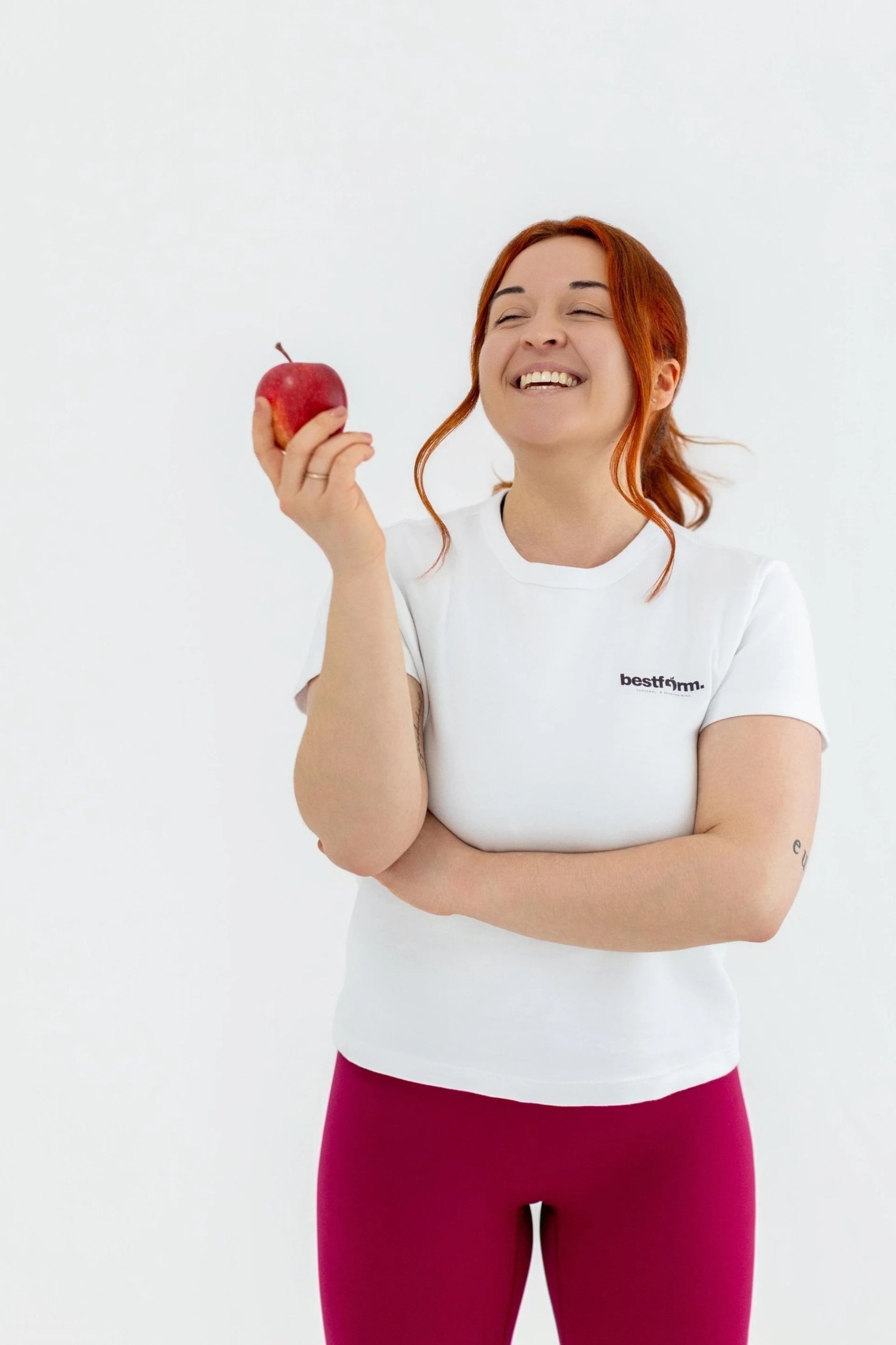 Junge Frau mit roten Haaren hält einen roten Apfel, trägt ein weißes T-Shirt mit dem Logo "bestform" und rote Leggings, lächelnd auf einem weißen Hintergrund.