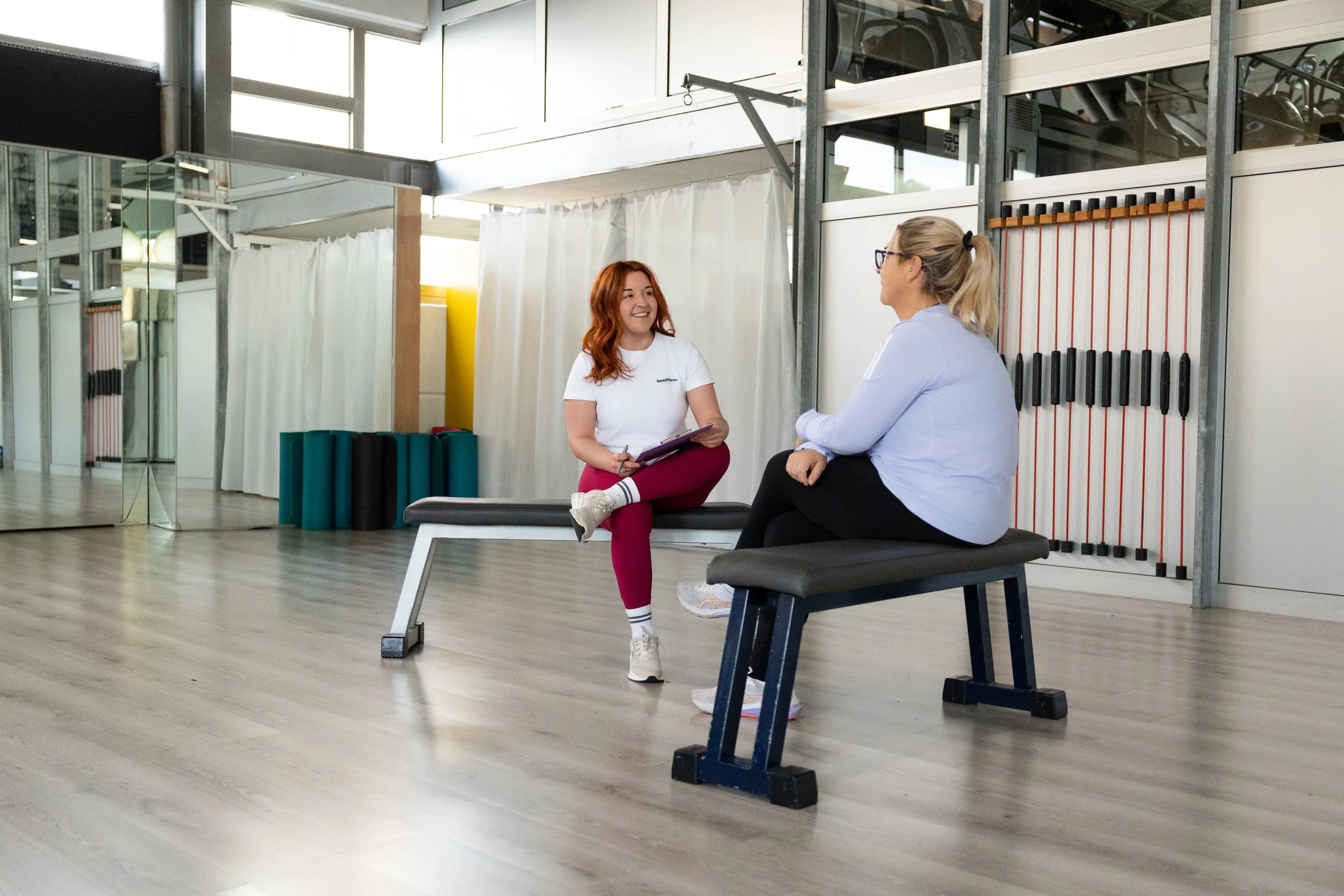 Zwei Frauen sitzen in einem modernen Fitnessstudio und führen ein Gespräch. Eine Frau trägt sportliche Kleidung mit einem weißen T-Shirt und rot/pinkfarbenen Leggings, die andere trägt eine weiße Jacke und schwarze Leggings. Der Raum hat große Fenster und eine Spiegelwand.