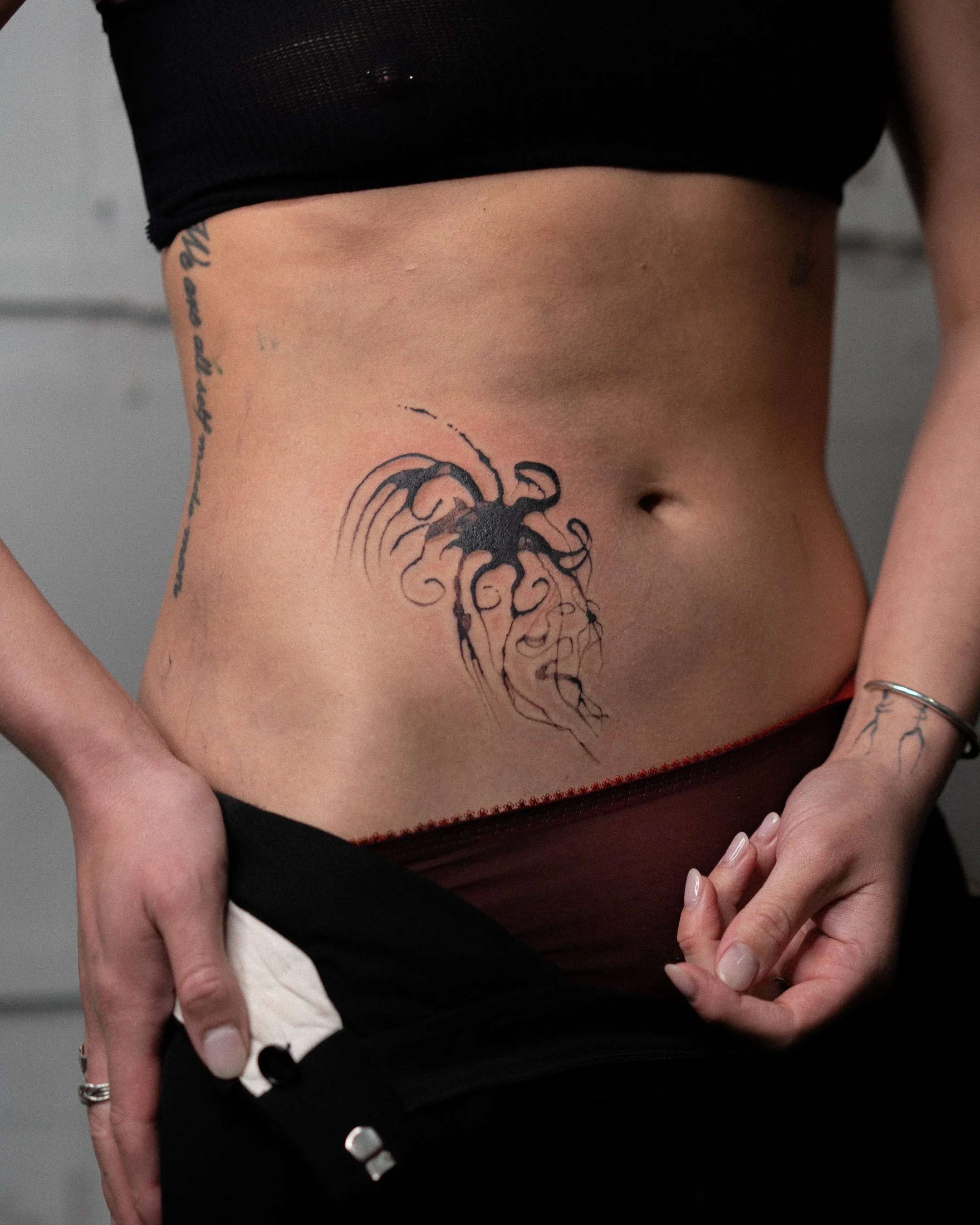 Ventre d'une personne avec tatouage d'une pieuvre sur le côté, portant un haut noir et des bijoux aux doigts.