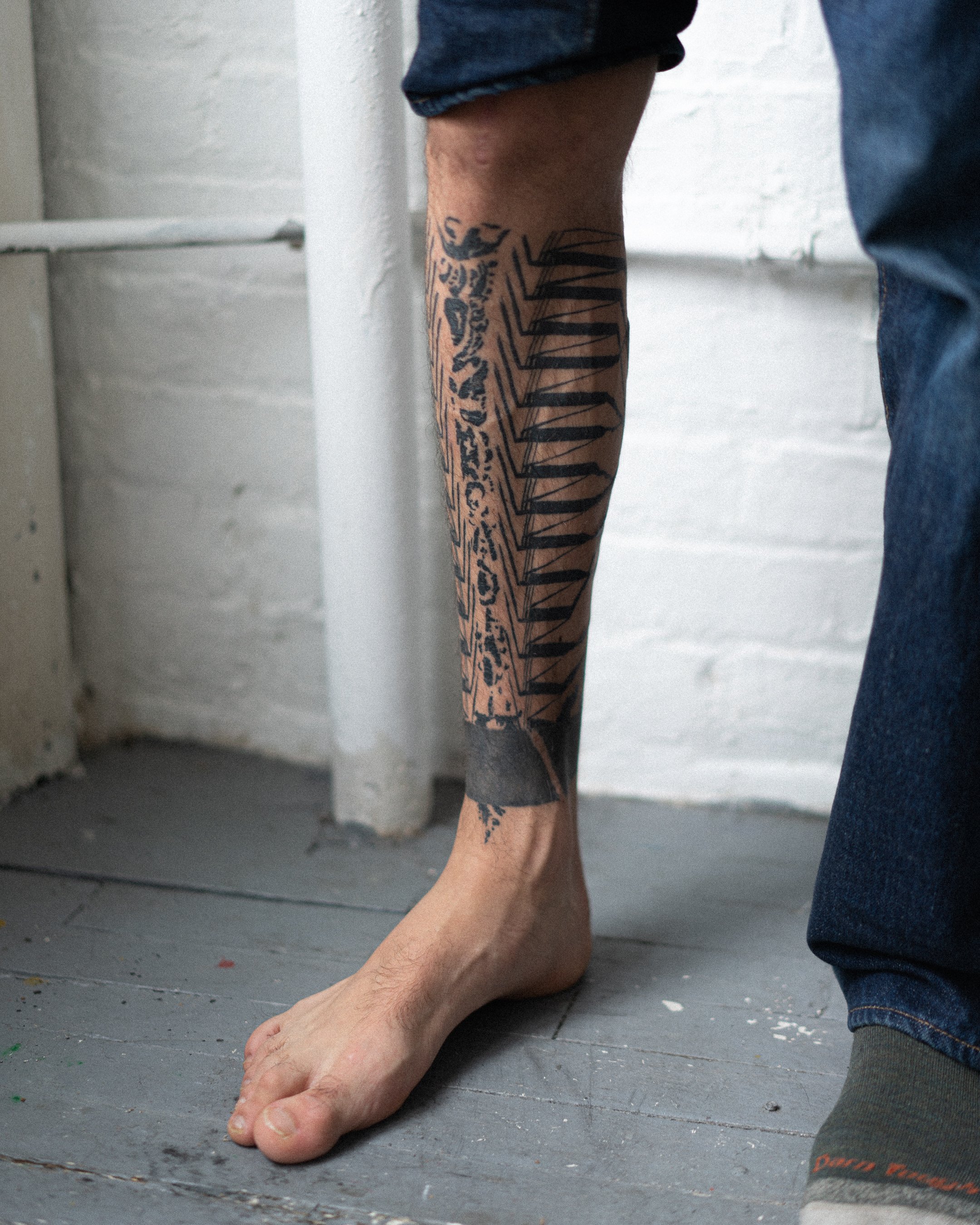 Un pied humain à plat sur un sol gris, avec un tatouage au bras visible et une personne en jean à côté.