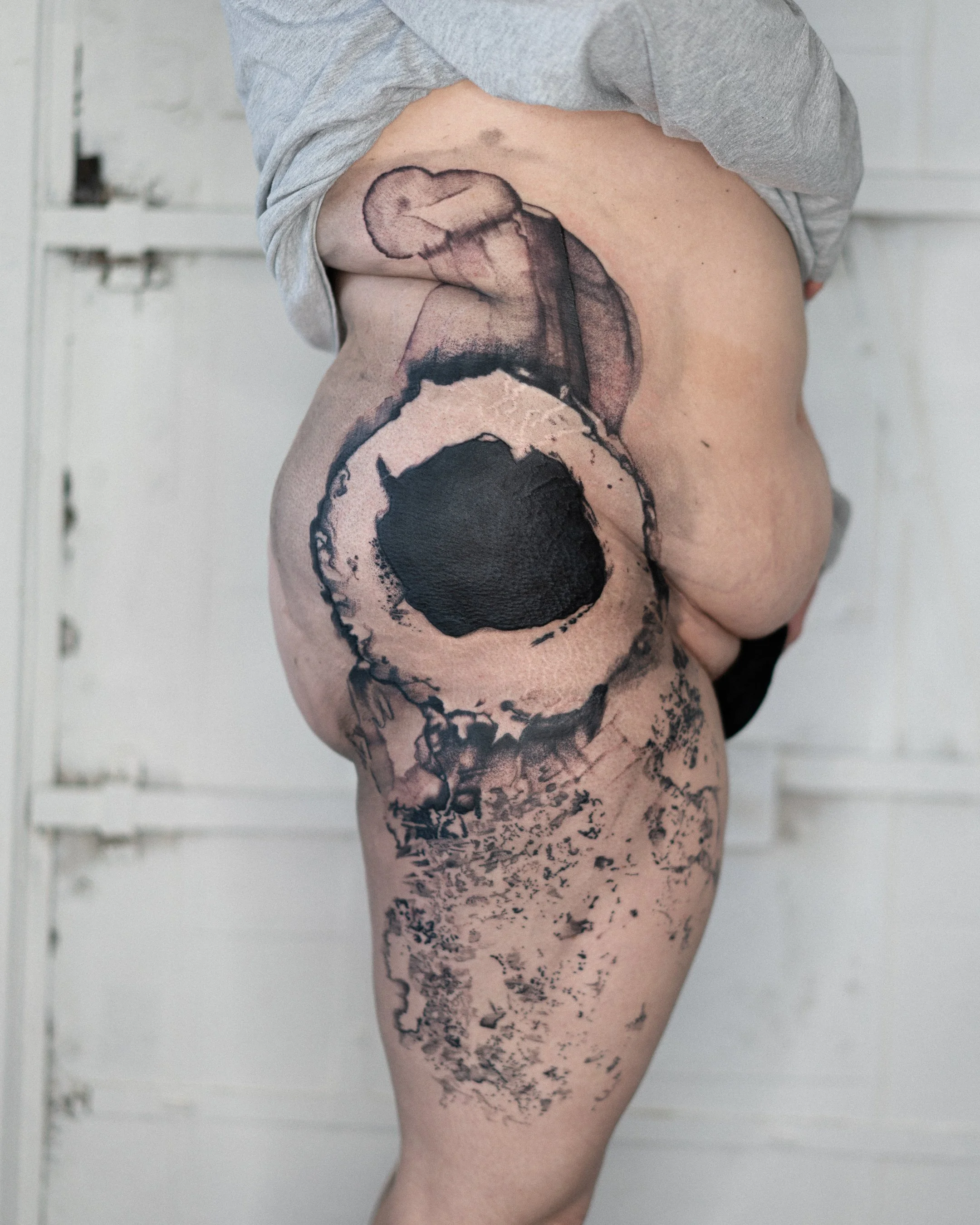 Tatouage abstractionnel noir sur la cuisse et le genou d'une personne nue, contre un mur blanc.