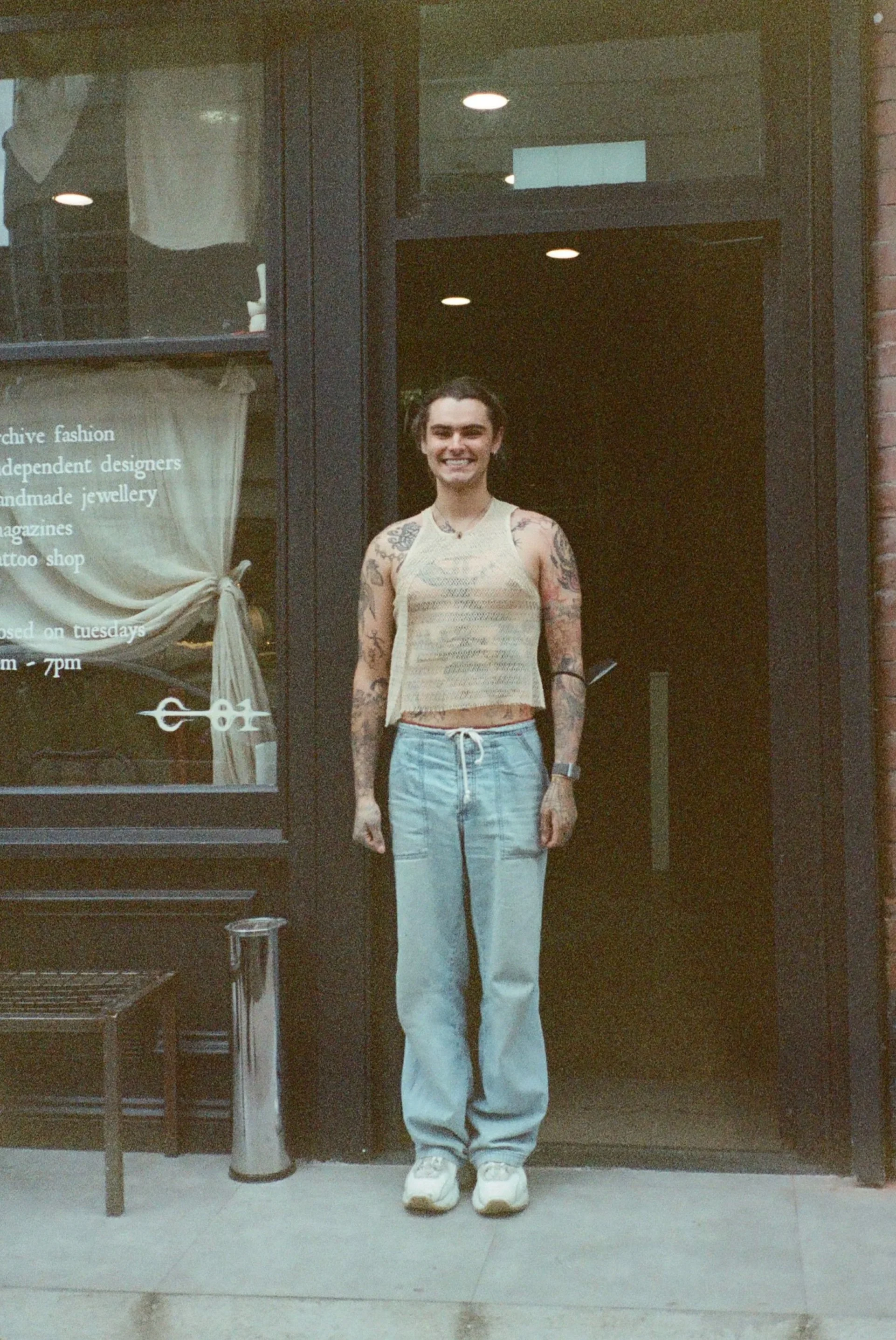 Jeune femme avec tatouages, souriante, portant un débardeur beige et un pantalon blanc, debout devant une porte en verre avec notes en français, à l'intérieur d'un bâtiment.