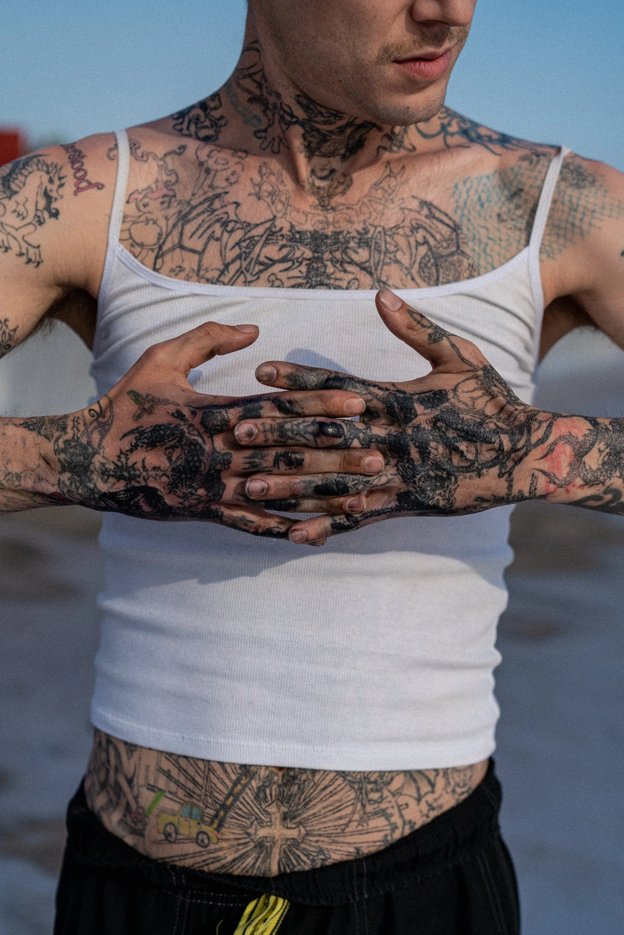 Un homme avec de nombreux tatouages sur le torse, les bras et l'abdomen, portant un débardeur blanc et des pantalons noirs. La personne semble tenir sa main sur son torse et a des tatouages colorés et noirs visibles.