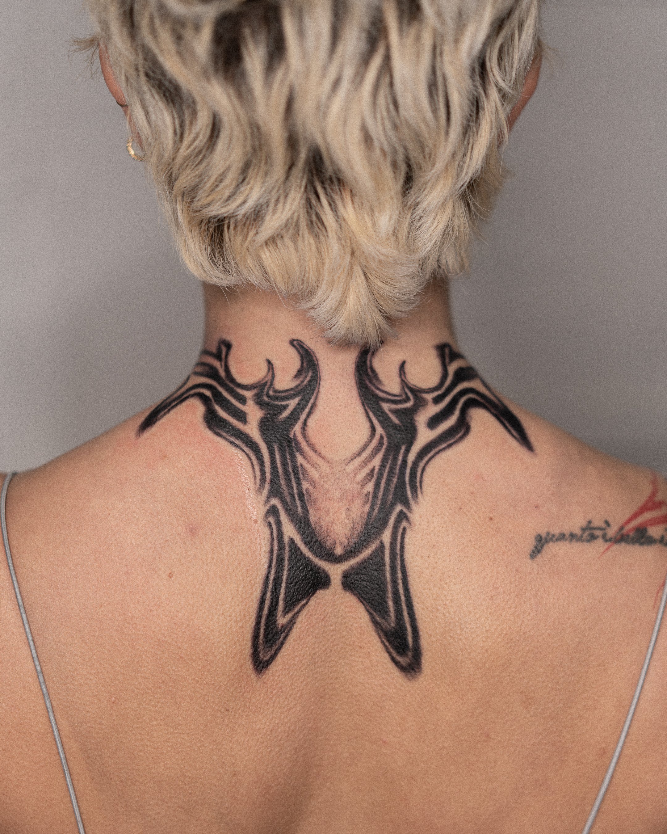 Une personne sous les cheveux courts et argentés porte un tatouage noir sur le dos du cou, représentant un animal ou un motif abstrait.