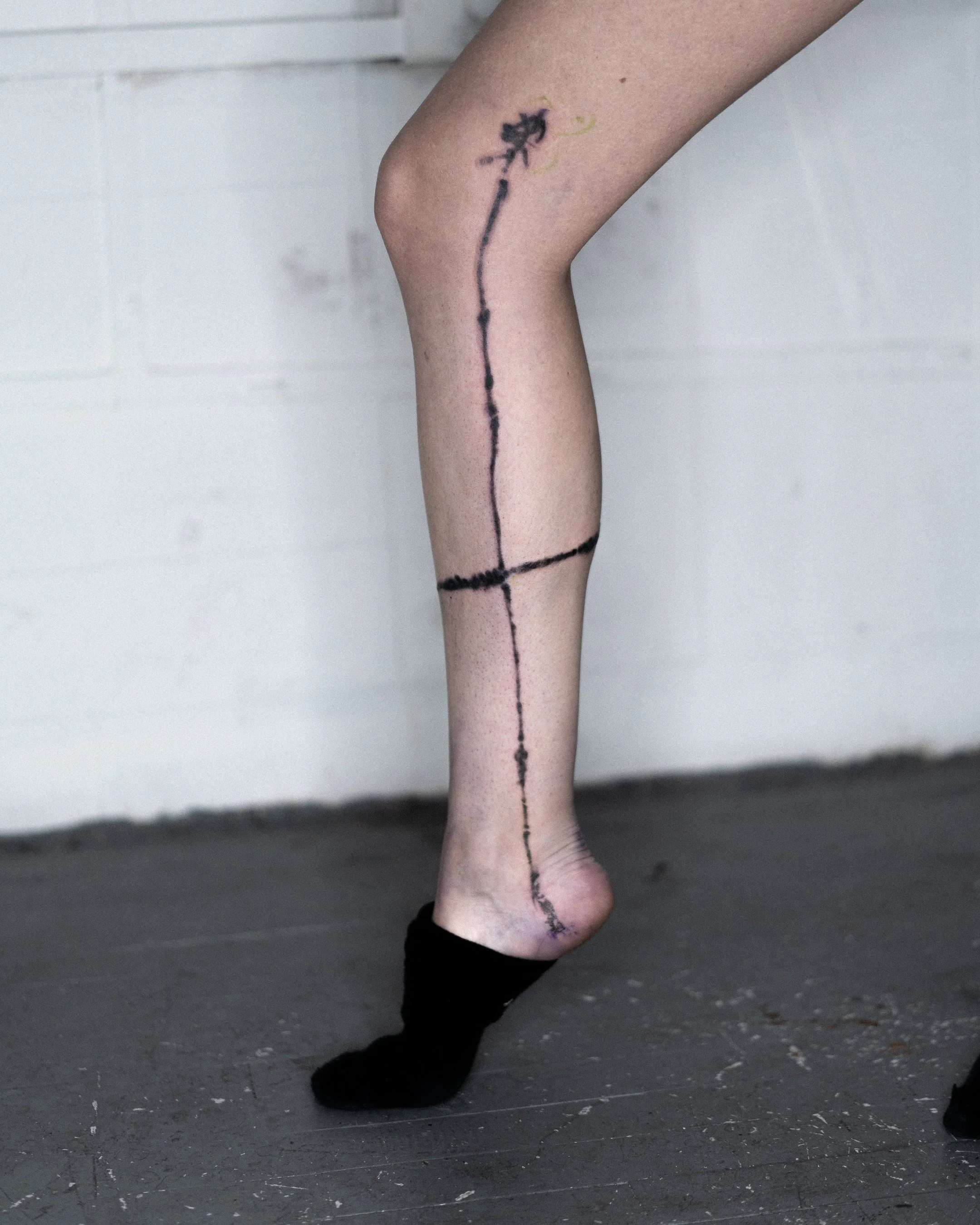 Une jambe de personne portant un collant noir, avec un tatouage de ligne noire représentant un skateboard et un dragon. La personne porte une chaussette noire, et l'arrière-plan est une mur blanc avec un sol en béton noir.