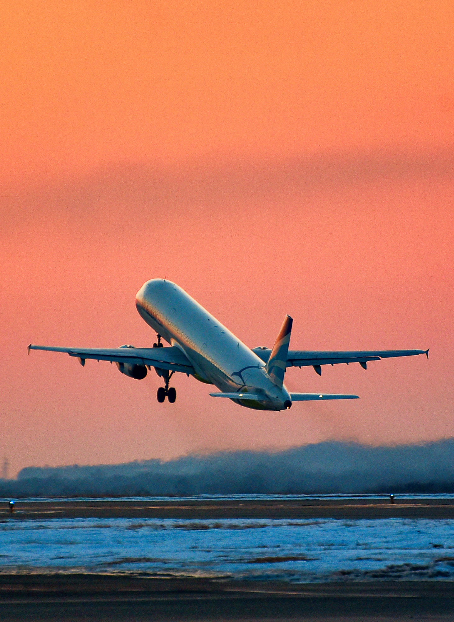 A commercial airplane is taking off or landing on a runway near the ocean during sunset, with a pinkish-orange sky and mountains in the background.