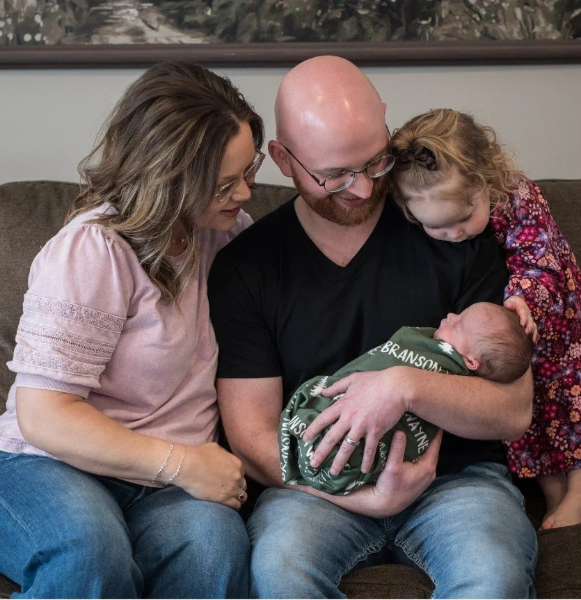 Family of four with parents and two children looking at their newborn, sitting on a couch.