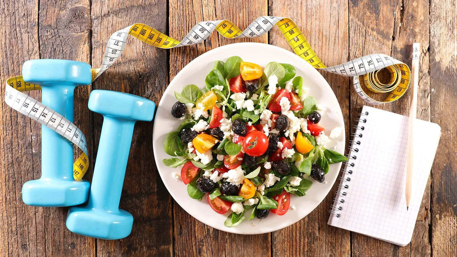 A plate of fresh vegetable salad with cherry tomatoes, black olives, feta cheese, and greens, alongside light blue dumbbells, a yellow and white measuring tape, a notepad, and a pencil on a rustic wooden surface.