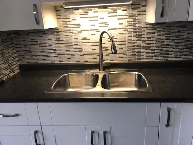 Kitchen sink with a black countertop, stainless steel double basin sink, and a chrome faucet, with a mosaic tile backsplash and white cabinets.