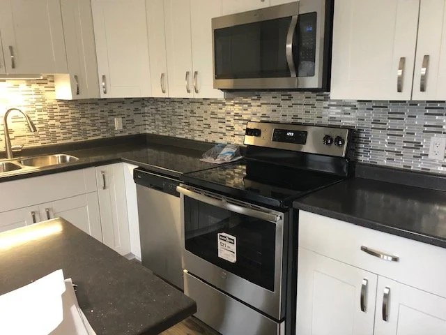 Modern kitchen with white cabinets, black countertops, stainless steel microwave, oven, and dishwasher, glass tile backsplash, and a stainless steel sink.
