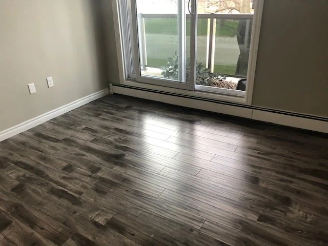 Empty room with wood-look laminate flooring, beige walls, white baseboards, two electrical outlets, and a sliding glass door leading outside.