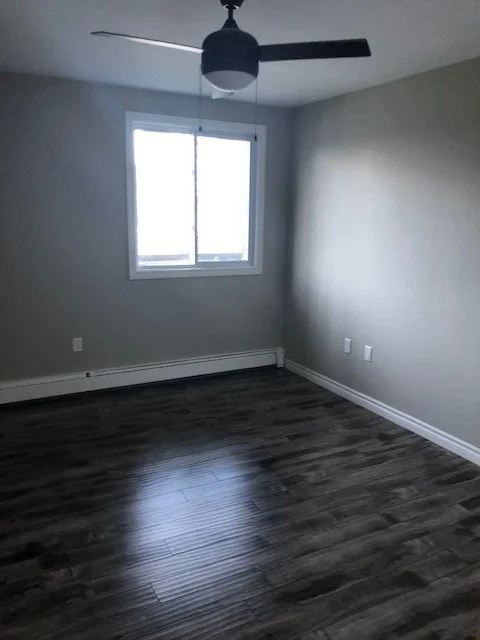 Empty room with gray walls, dark wood flooring, a window, a ceiling fan, and electrical outlets.
