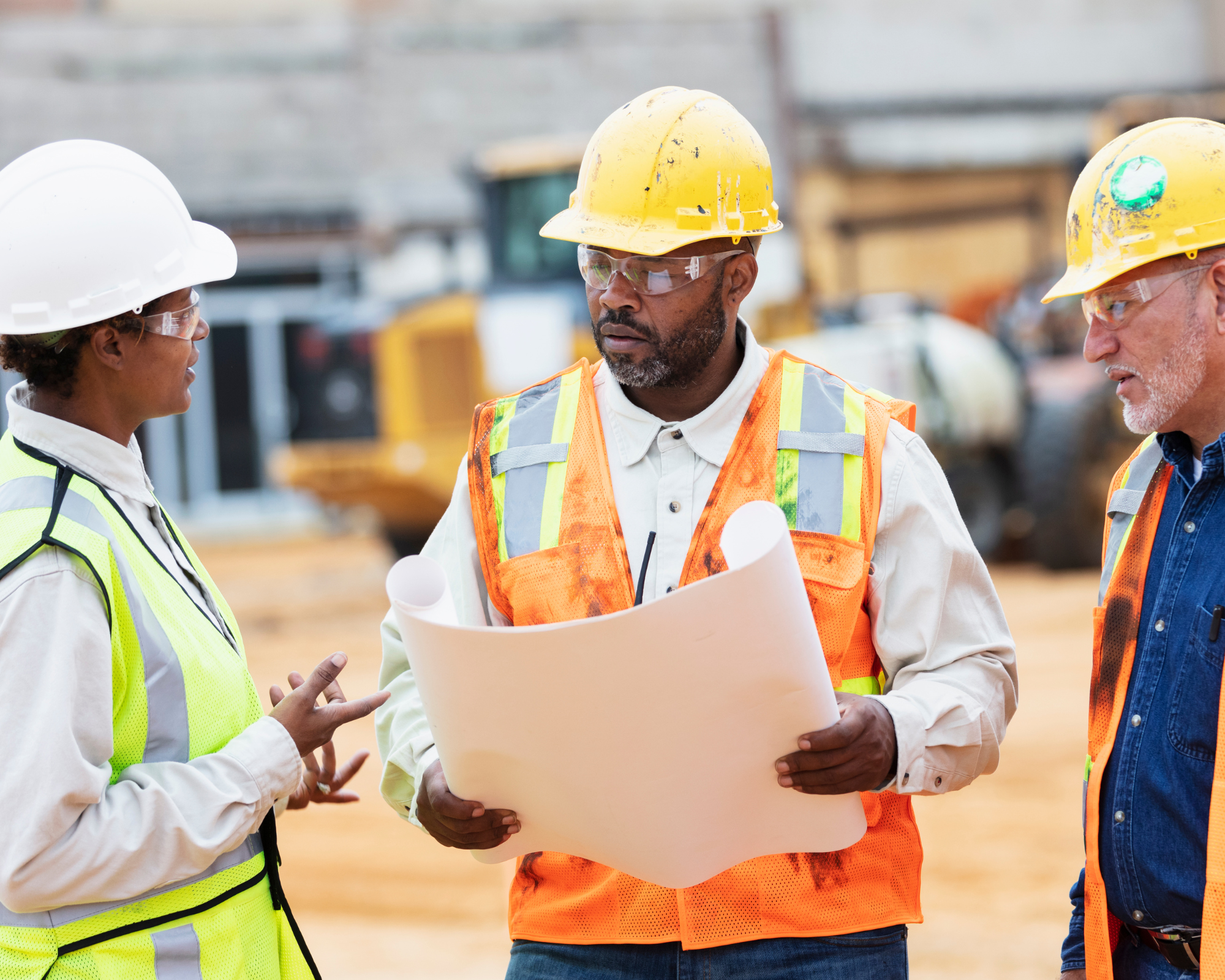 Three construction workers in safety helmets and vests having a discussion at a construction site, with construction equipment in the background.