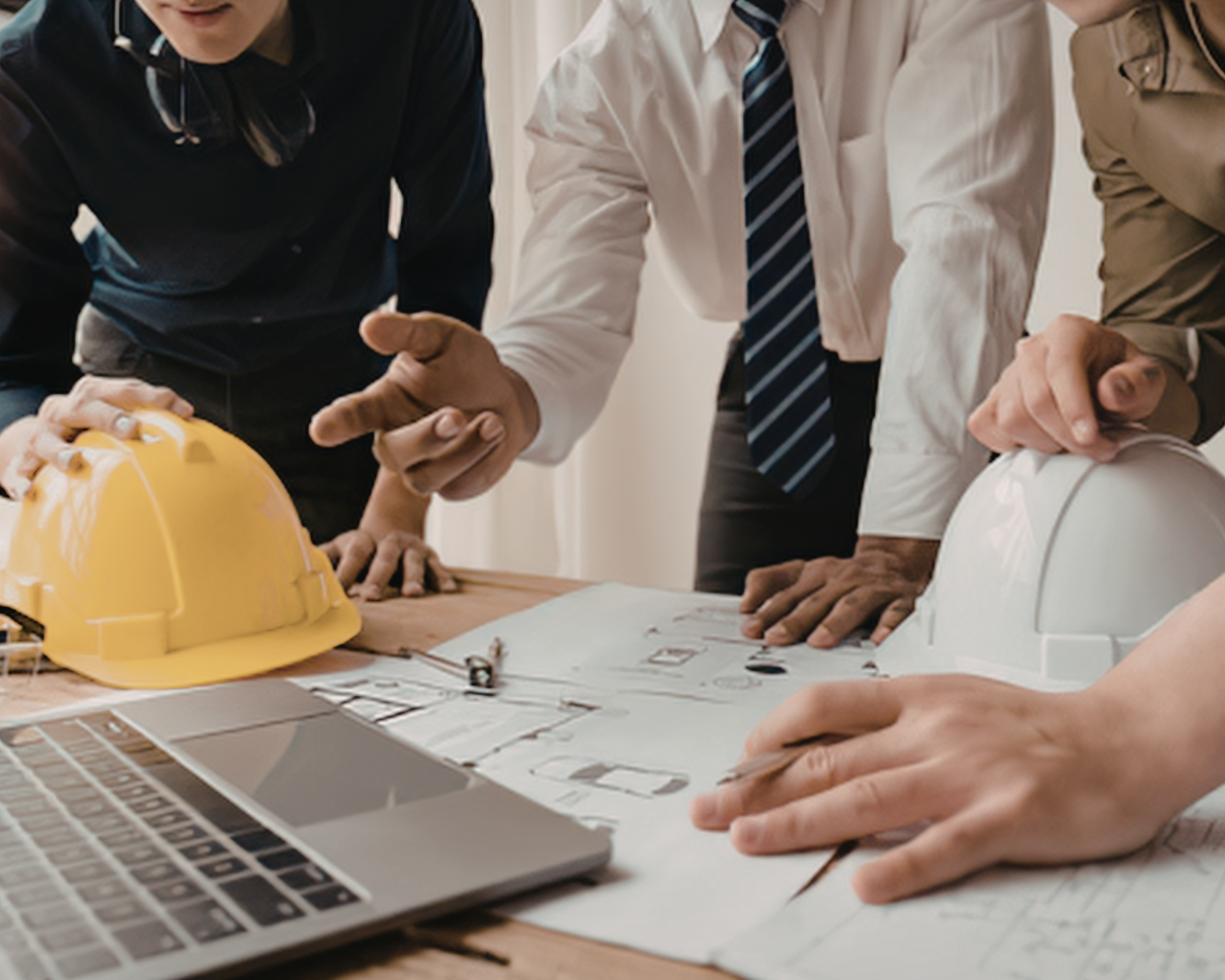 Group of people gathered around a desk with a yellow safety helmet, a laptop, and blueprints, discussing a project.