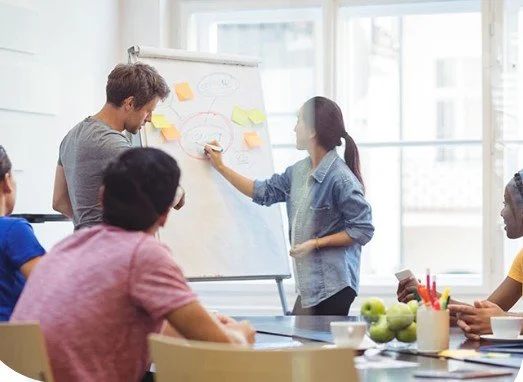 A woman presents at a whiteboard as a man points to it during a meeting with four seated colleagues in a bright office.