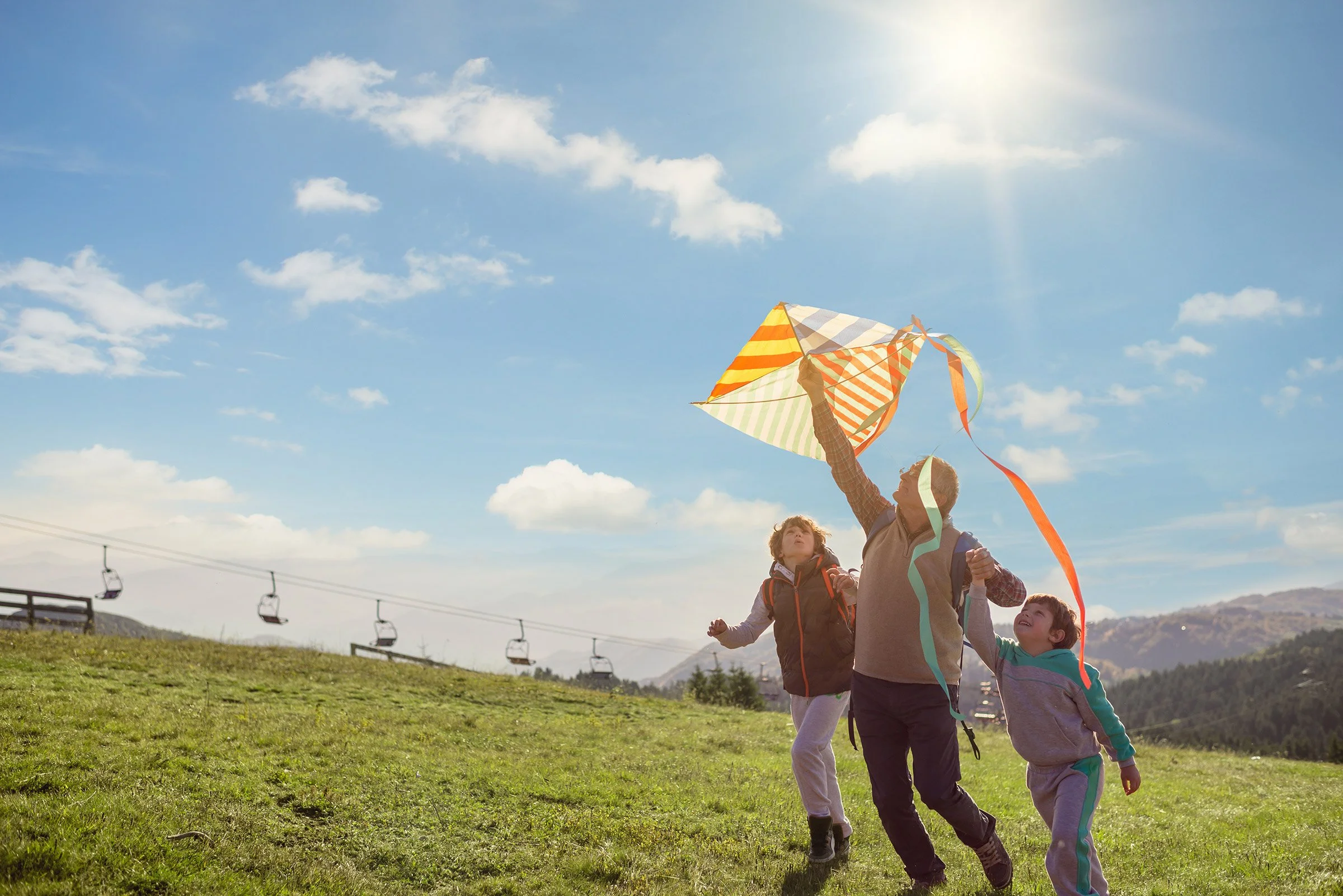 Niños y un adulto volando papalotes en un campo con colinas y sillas de telesilla en el fondo, cielo despejado con algunas nubes y sol brillante.