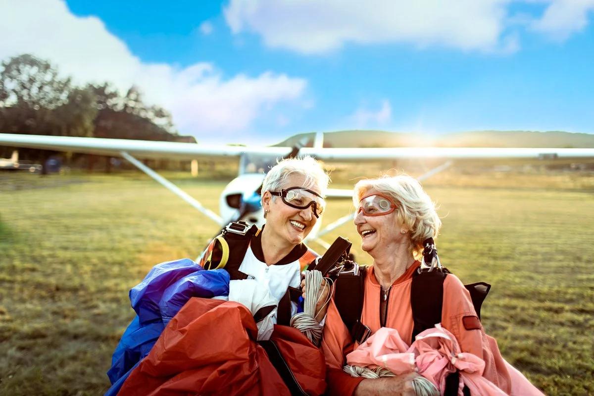 Dos personas mayores sonrientes con equipo de parapente y gafas de protección, en un campo con un avión pequeño al fondo, al atardecer.