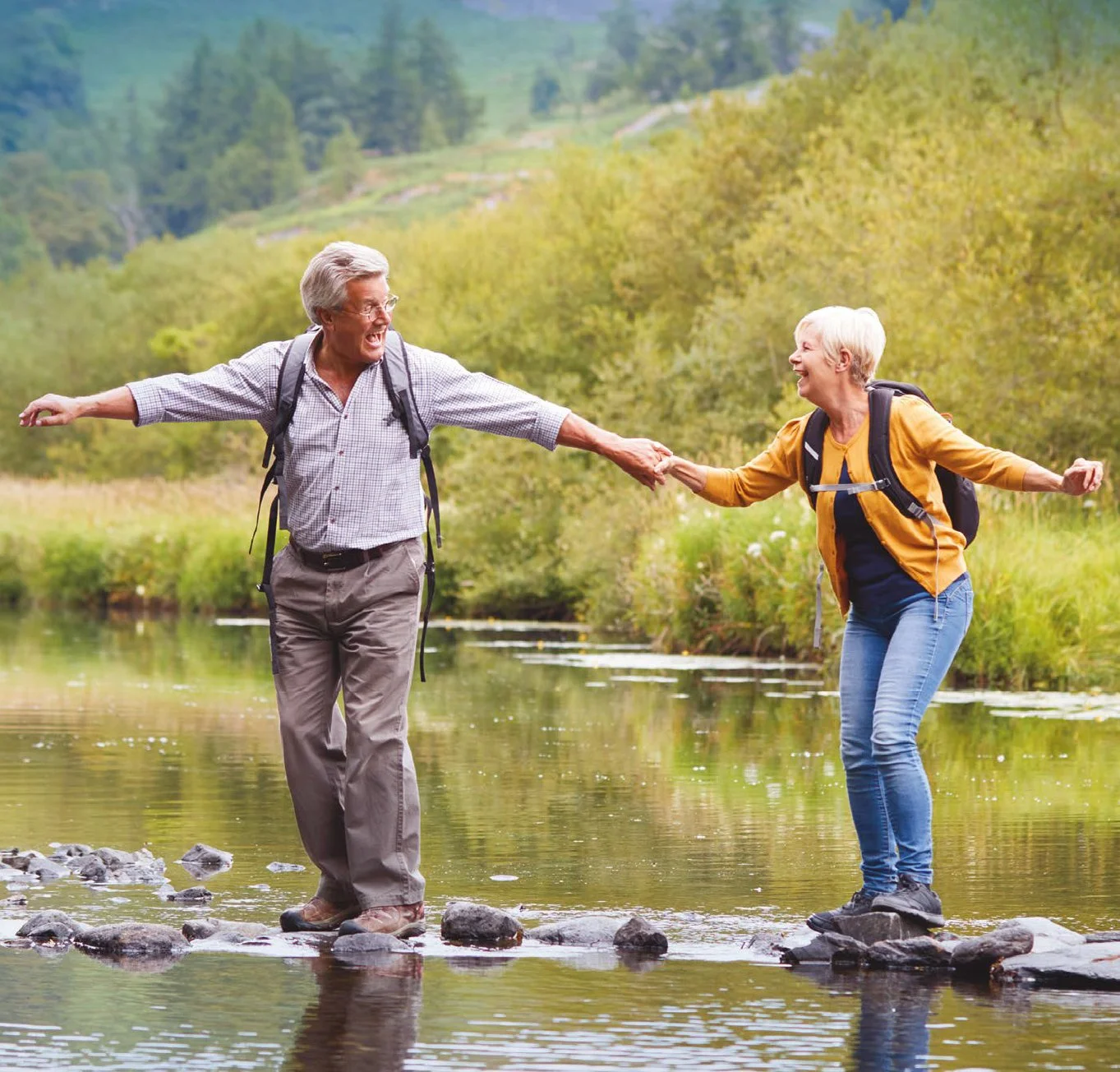 Dos adultos mayores, un hombre y una mujer con mochilas, jugando y sonriendo en un río rodeado de naturaleza y árboles en fondo.
