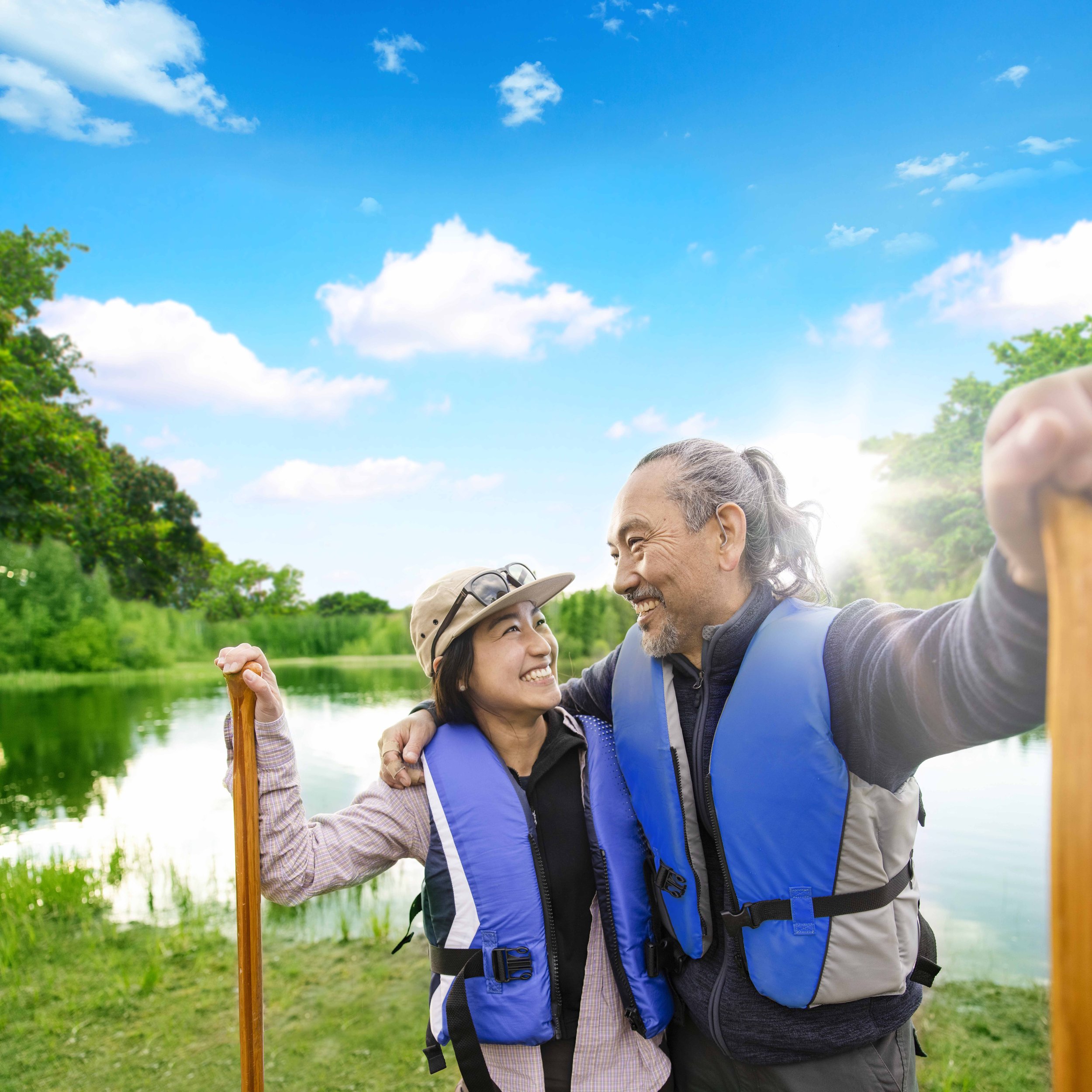 Dos personas mayores, un hombre y una mujer, sonriendo y abrazándose en un entorno natural junto a un río o lago, con árboles y un cielo azul con nubes en el fondo, en un día soleado.