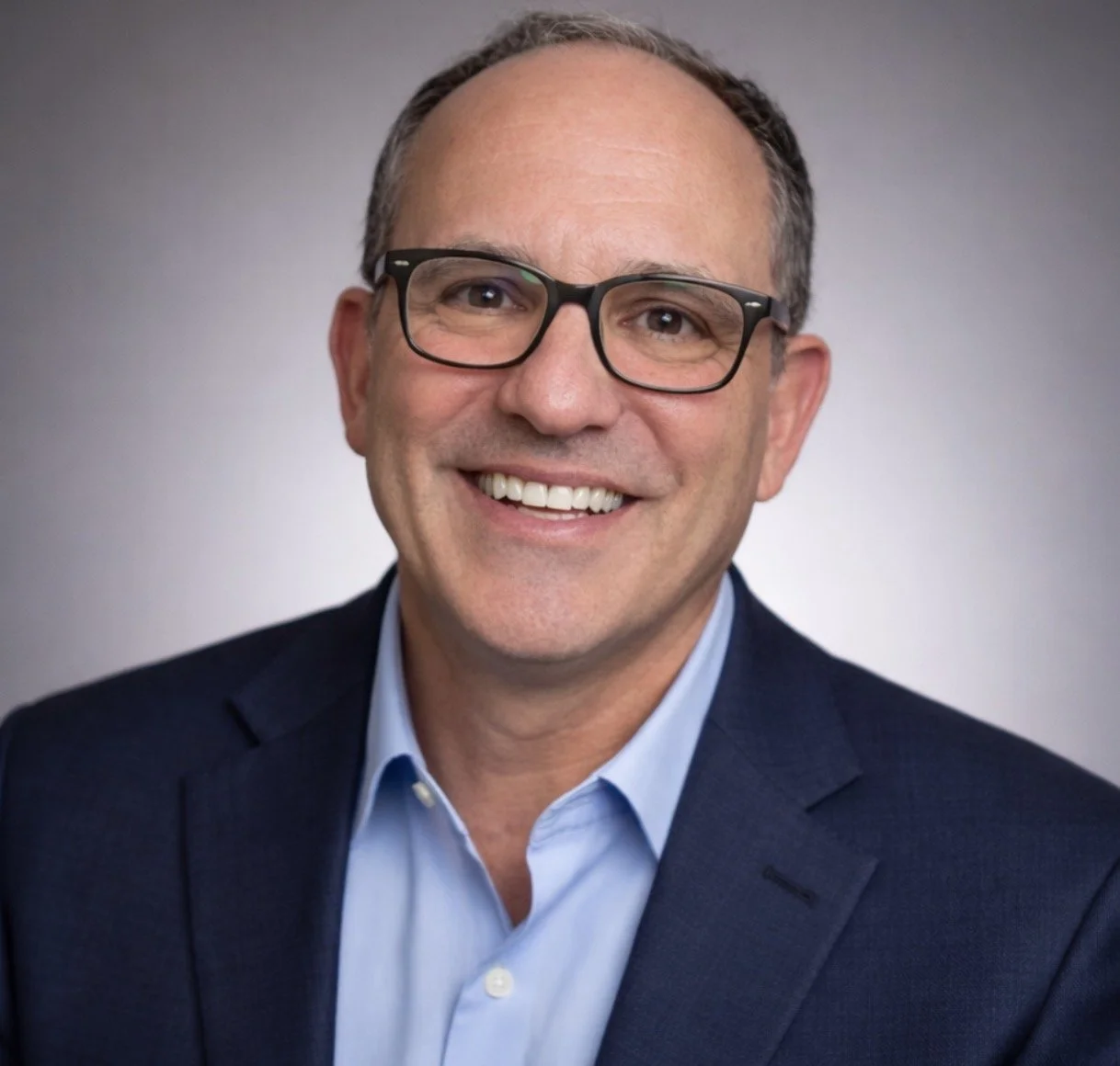 A man with glasses smiling, wearing a dark suit and light blue shirt, posed against a plain gray background.