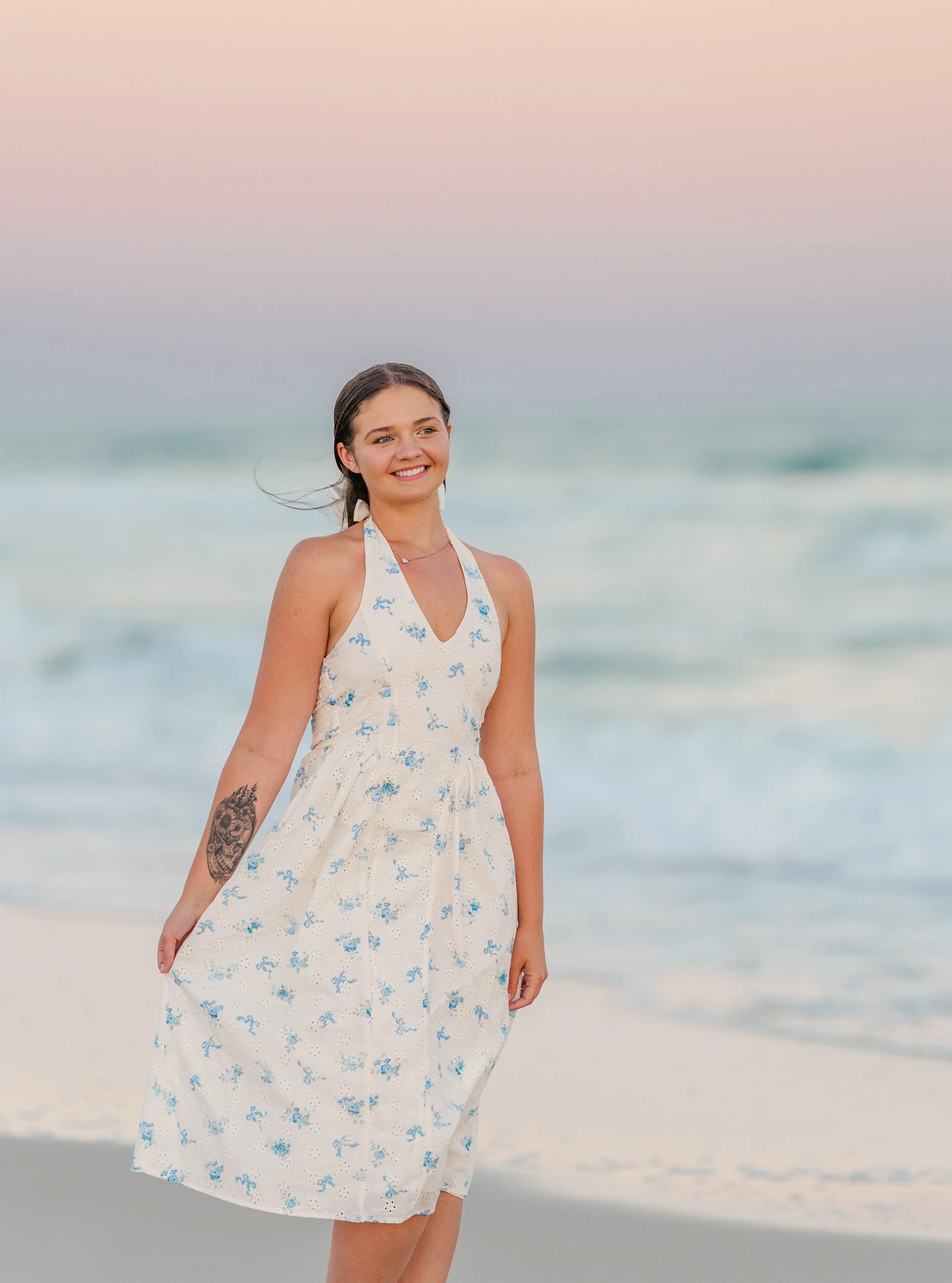 Young woman at the beach with long hair, wearing a white sundress with blue floral pattern, smiling with ocean waves in the background.