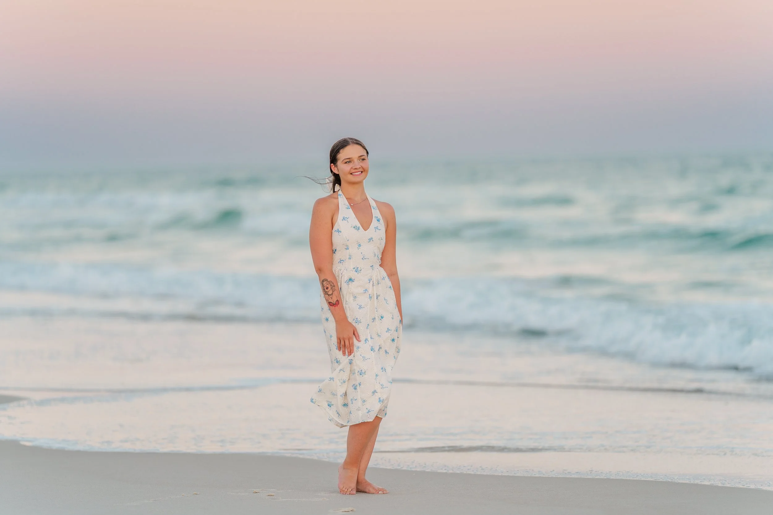 A woman standing barefoot on the beach with ocean waves in the background during sunset, wearing a white dress with a blue floral pattern and tattoos on her left arm, smiling and looking to her left.