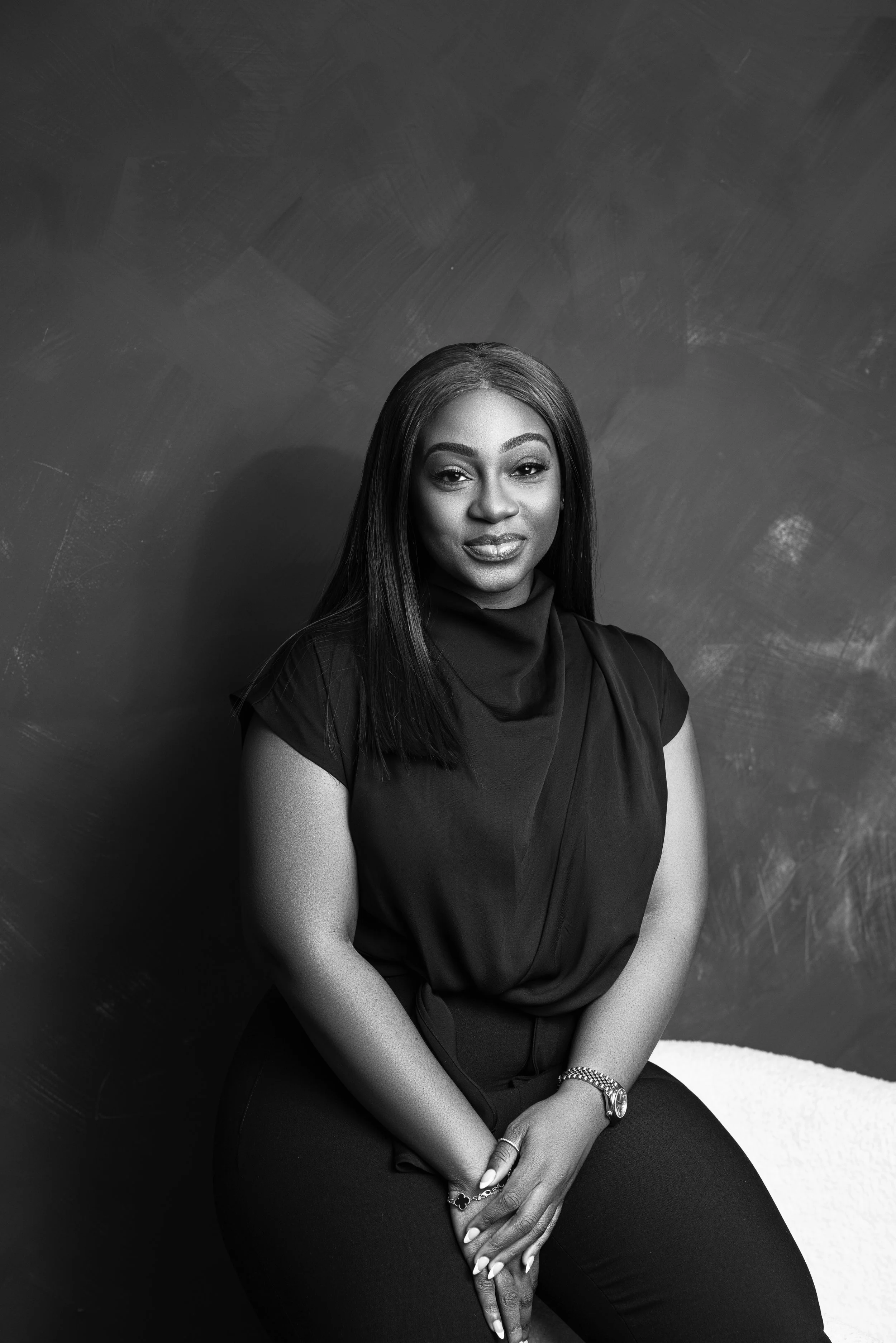 Black and white portrait of a woman with long straight hair, wearing a dark top with a high neckline, sitting against a textured wall with a subtle smile, hands clasped in front, wearing jewelry on her wrist and finger.