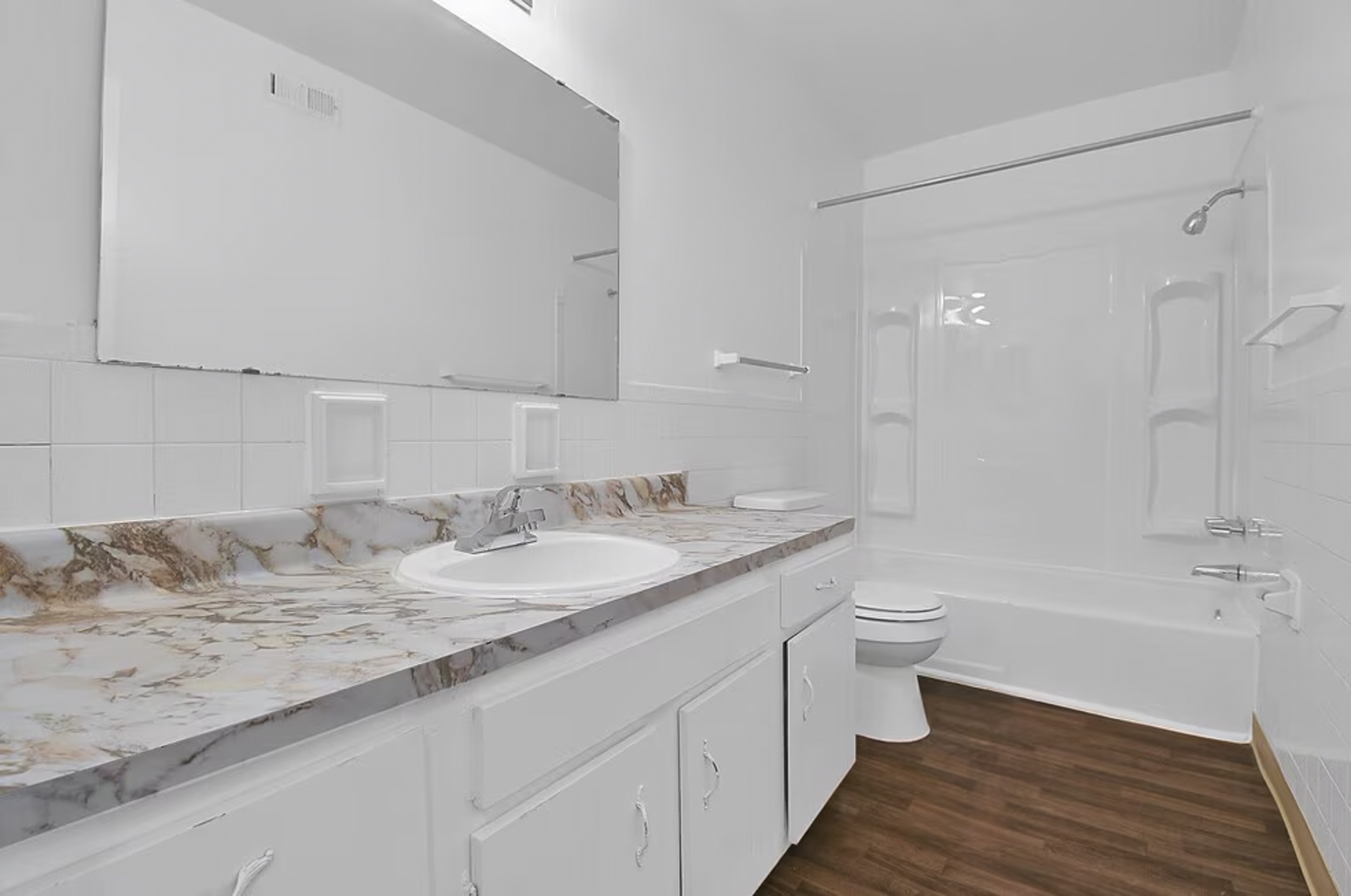 Bathroom with white walls, a marble countertop sink, a large mirror, a bathtub with a shower, a toilet, and hardwood flooring.