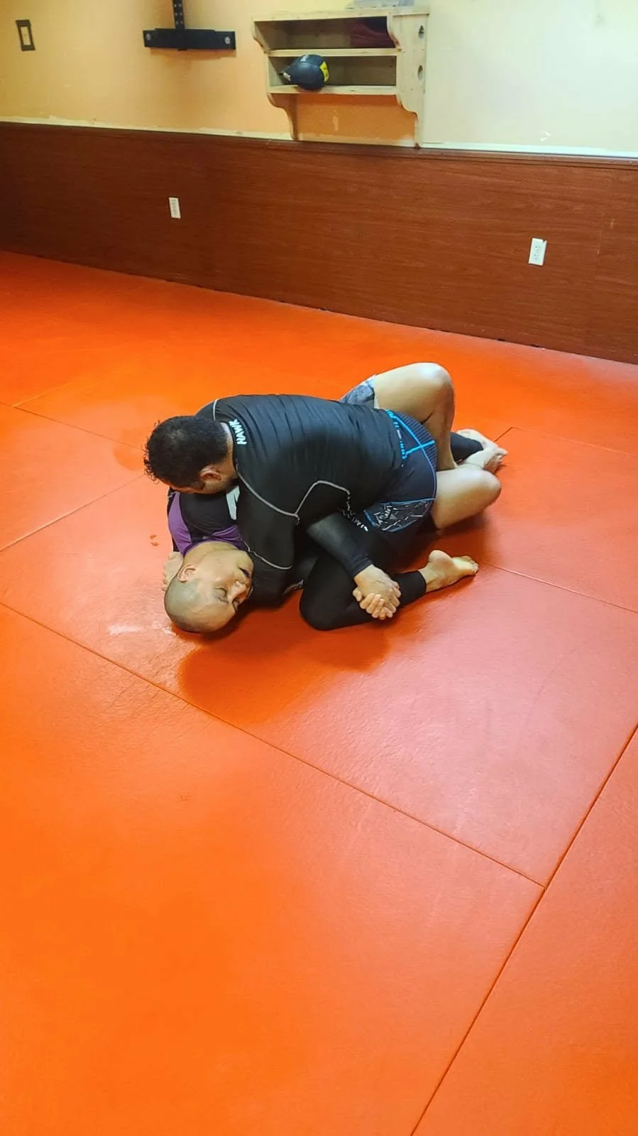 Two men practicing Brazilian Jiu-Jitsu on a red padded mat. One man is in a dominant position on top, while the other is on the ground.