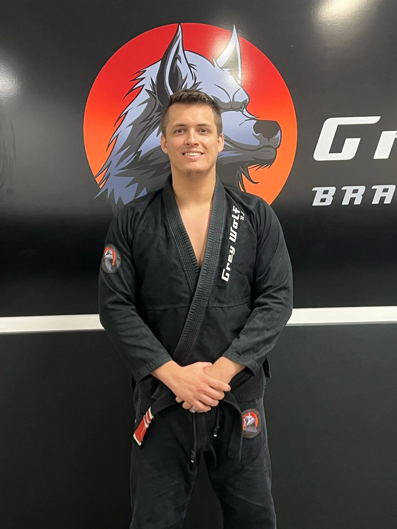 A smiling man in a black martial arts gi standing in front of a black wall with a logo of a wolf's head on an orange background. The man is wearing a black belt and has his hands clasped in front of him.