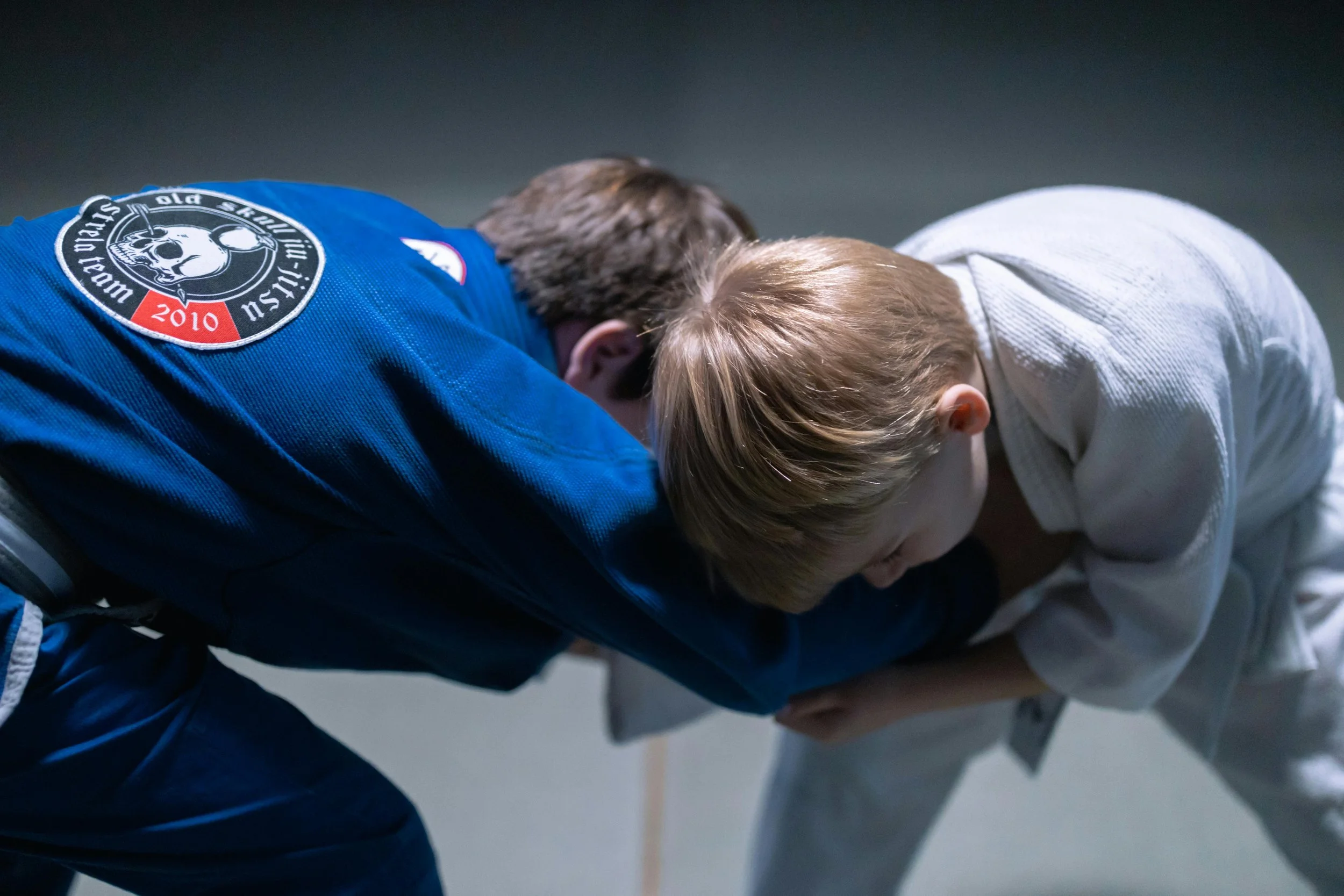 Two children, one in a blue judo gi with an emblem on the back, and the other in a white judo gi, are engaged in a judo match, leaning forward and gripping each other.