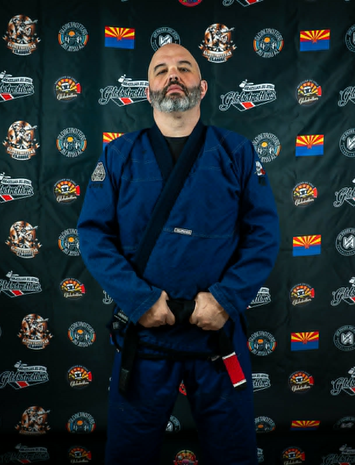 A man in a blue Brazilian Jiu-Jitsu gi with a black belt and red and white stripes stands with his hands clenched in front of him, against a backdrop featuring martial arts logos, the Arizona state flag, and other related symbols.