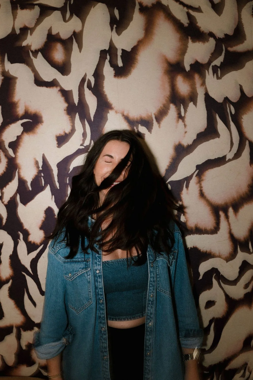 A woman with long dark hair smiling and covering her face partially with her hair, standing in front of a large floral mural.