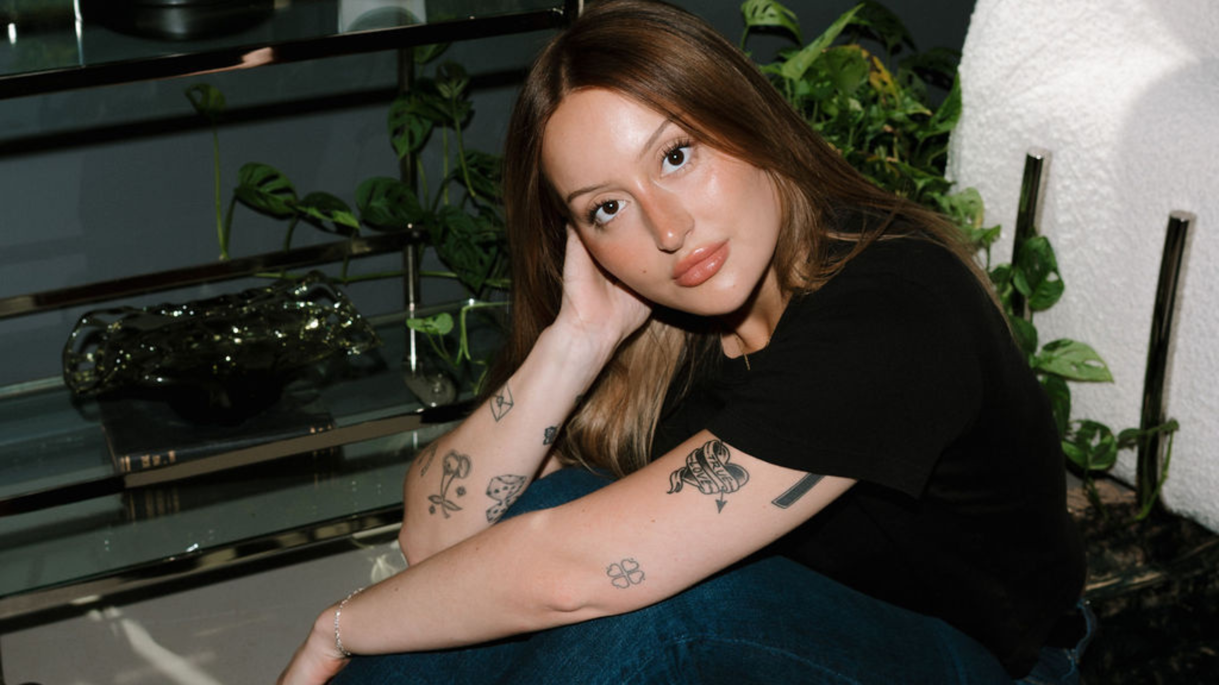Young woman with tattoos resting her head on her hand, sitting in a room with green plants and a glass shelf with decorative objects.