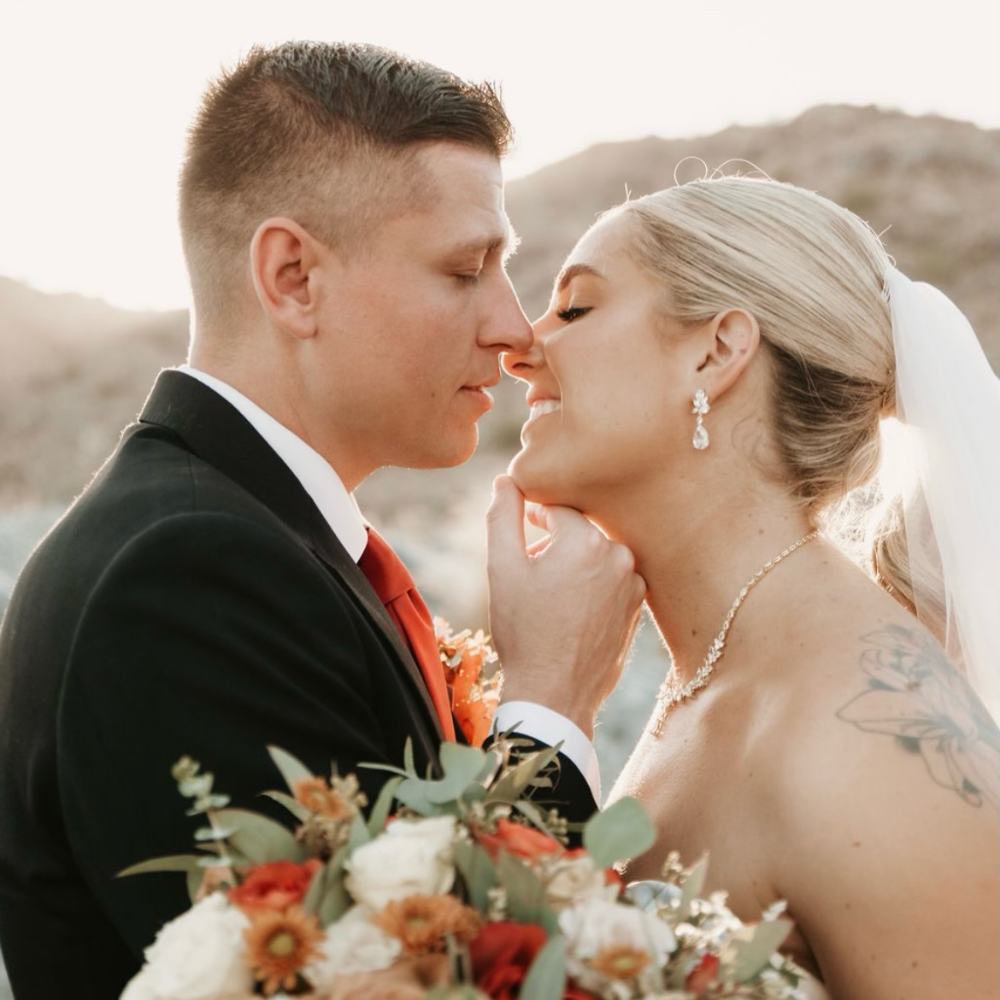 Bride and groom embracing outdoors, holding a bouquet with white and orange flowers, wearing wedding attire, against a natural background.