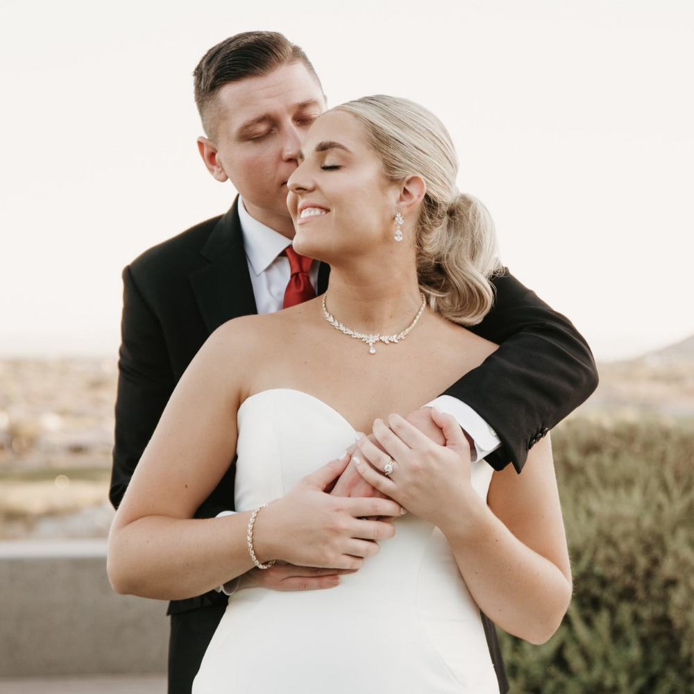 Bride and groom in formal attire embracing outdoors, with the groom kissing the bride's head, both smiling and appearing happy.