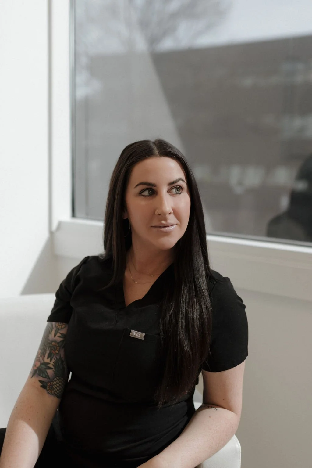 A woman with long dark hair sitting by a window, wearing a black shirt and displaying a colorful tattoo on her arm.