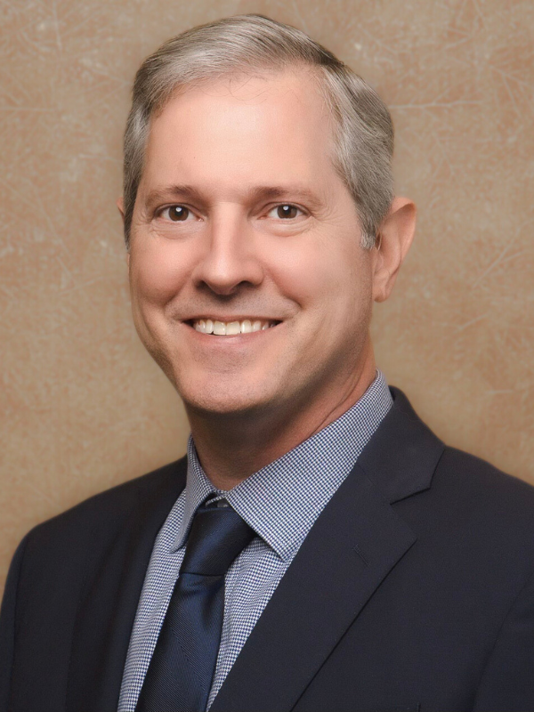 Portrait of a smiling man in a suit with a light gray background.